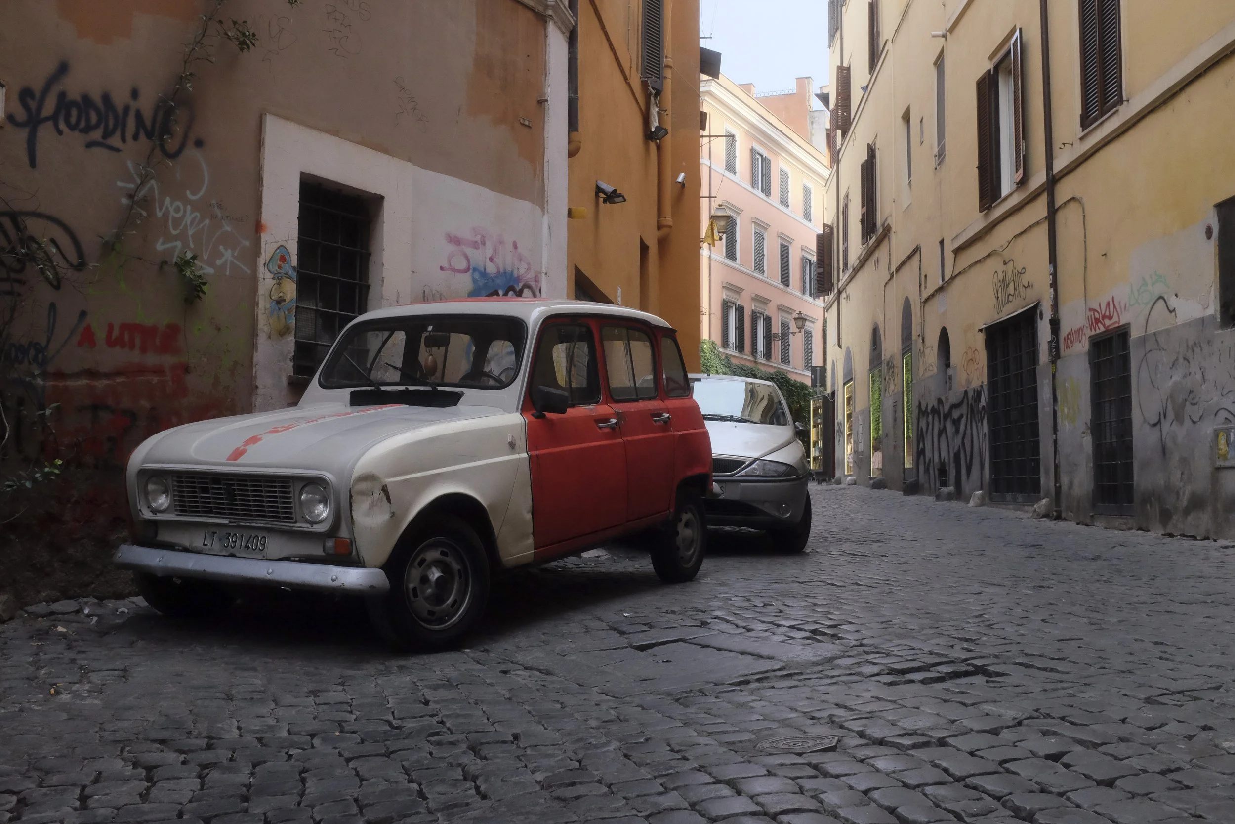 Old European street scene with narrow cobblestone street, parked vintage car, and multi-story buildings with shuttered windows and graffiti walls.