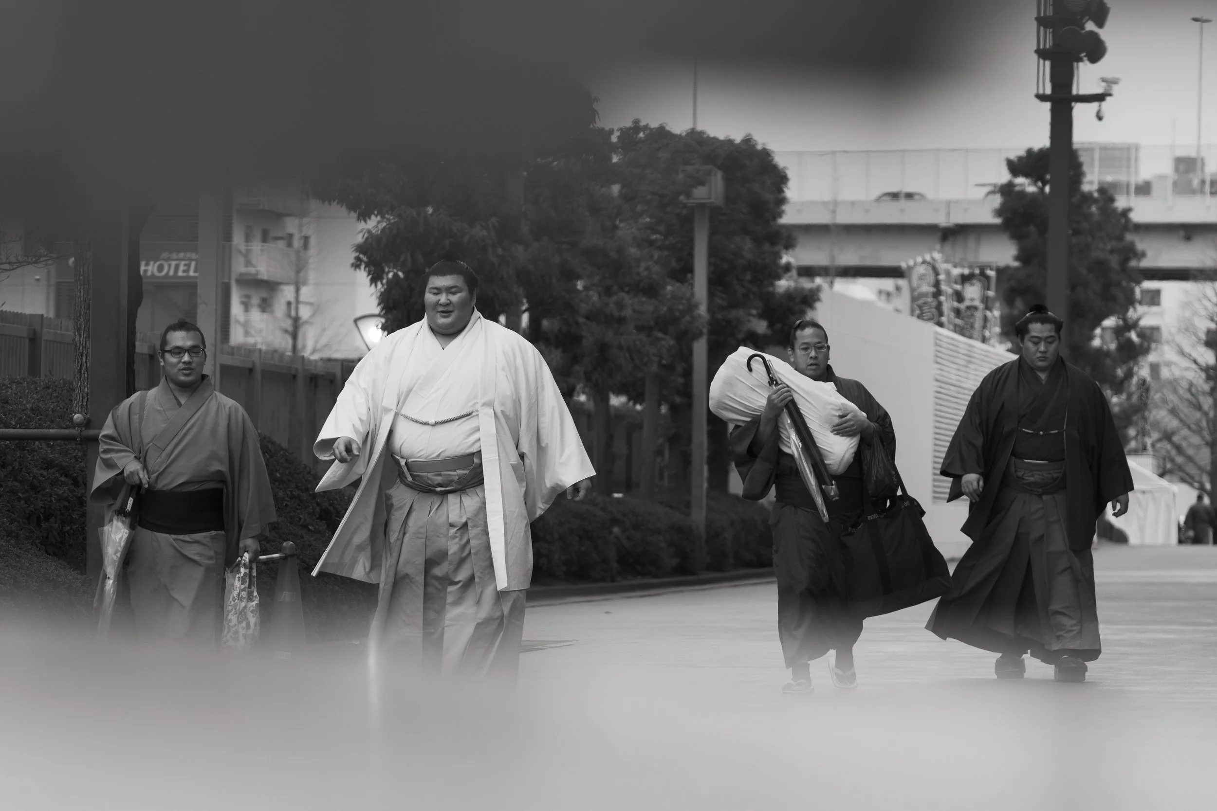 A group of four dressed in traditional Japanese garments, walking outdoors in a park or urban area, with trees, a fence, and a highway in the background, while one person carries an umbrella.