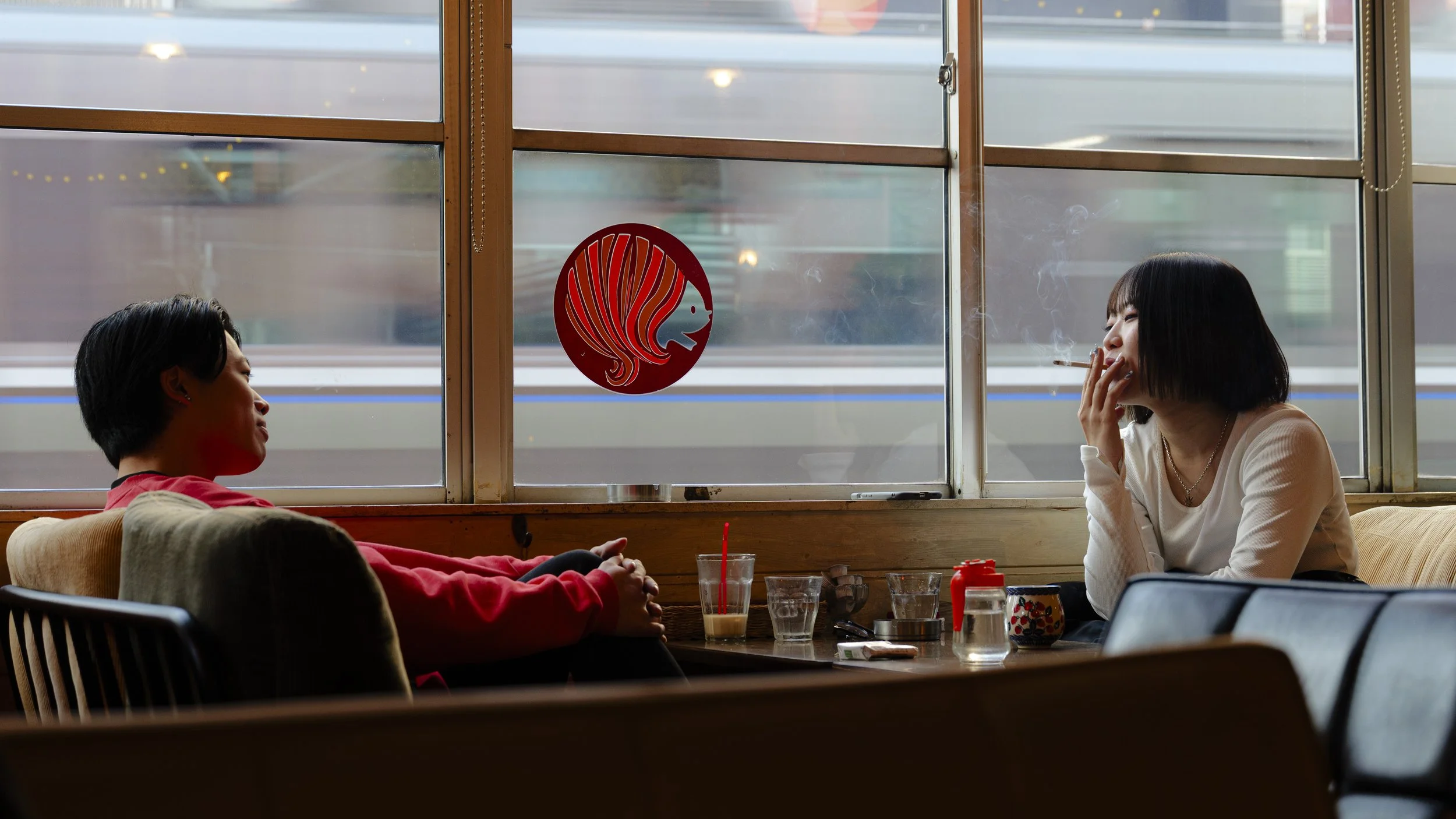 Two women sitting across from each other in a cafe by a window, with one smoking and the other talking, with a train visible outside.