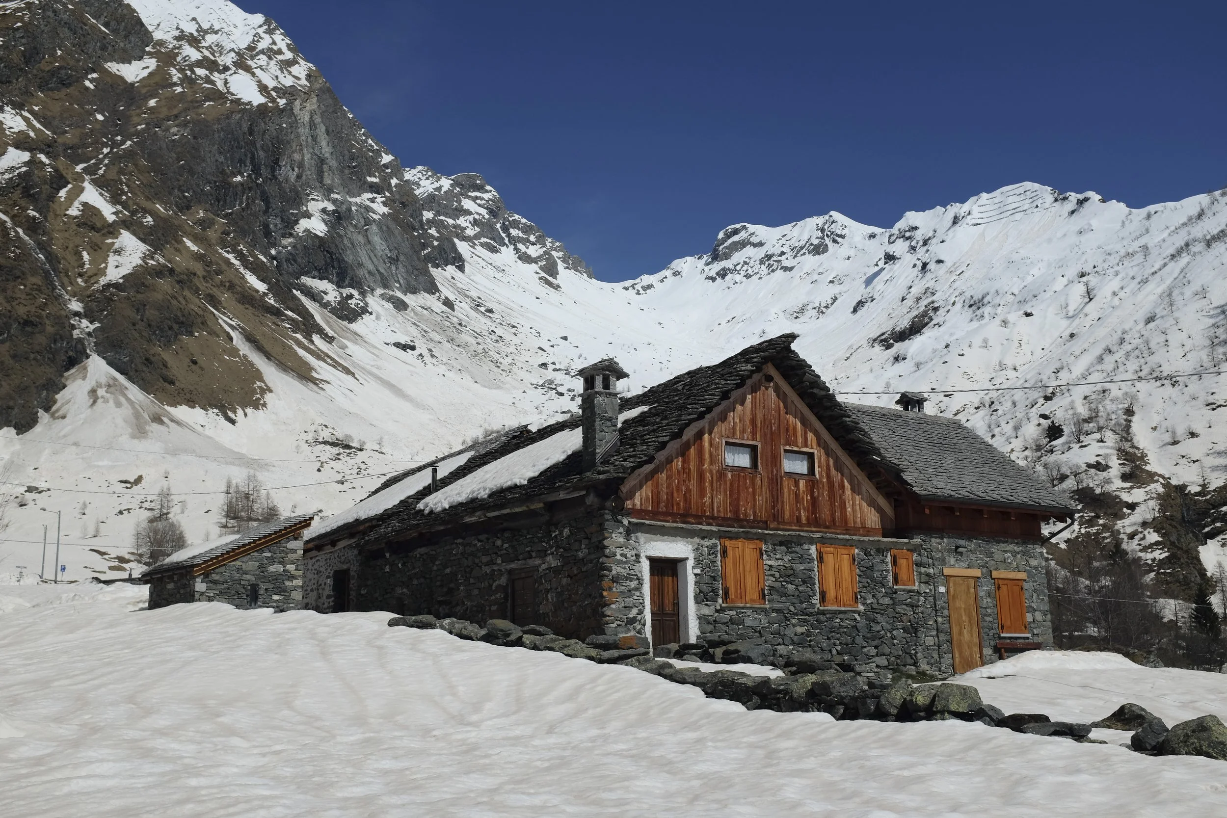 A rustic wooden house surrounded by snow, set against a backdrop of snow-capped mountain peaks under a clear blue sky.