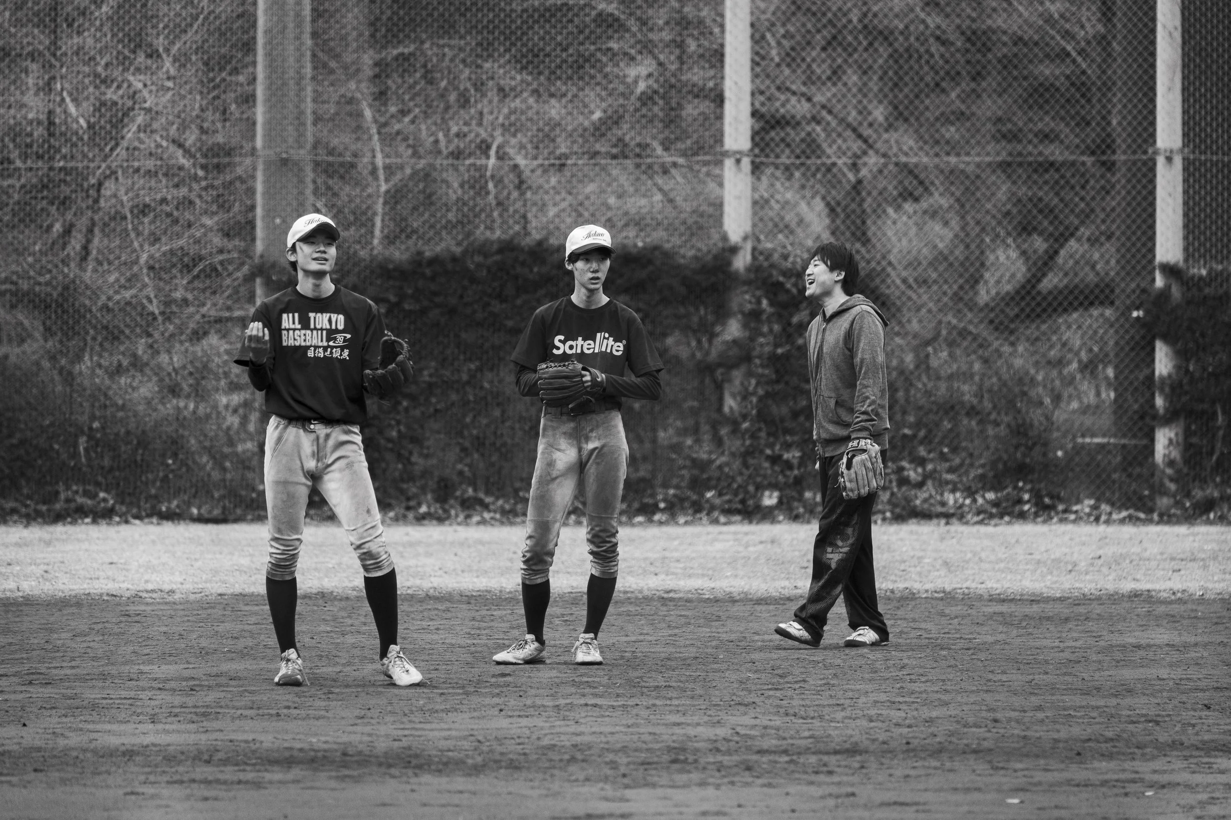 Three young baseball players and a young woman conversing on a baseball field. Two boys in baseball uniforms and a girl in casual clothes, with a chain-link fence and trees in the background.