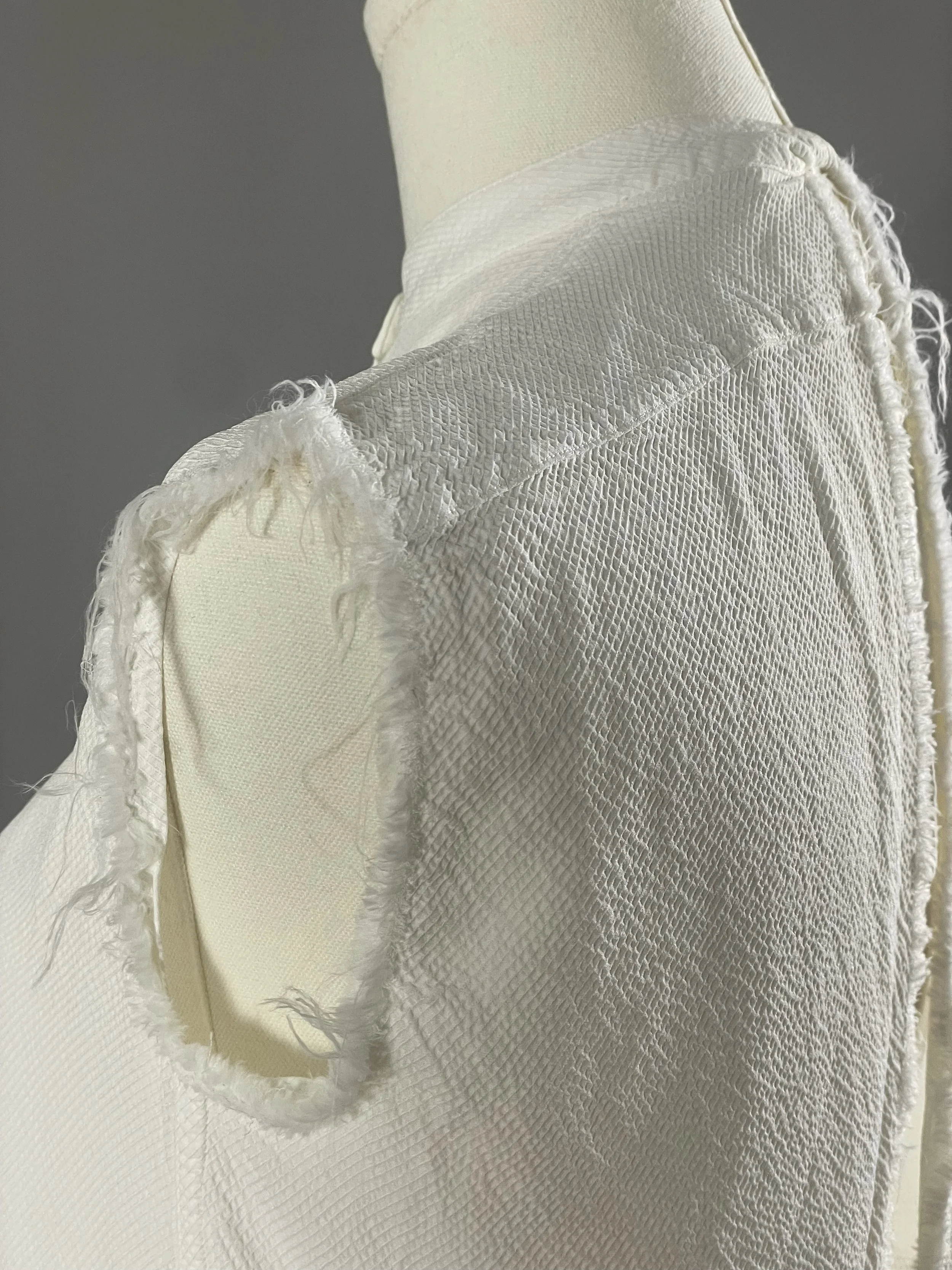 Detailed view of a textured white Helmut Lang garment on a dress form, focusing on the raw, frayed fringe of the shoulder construction.