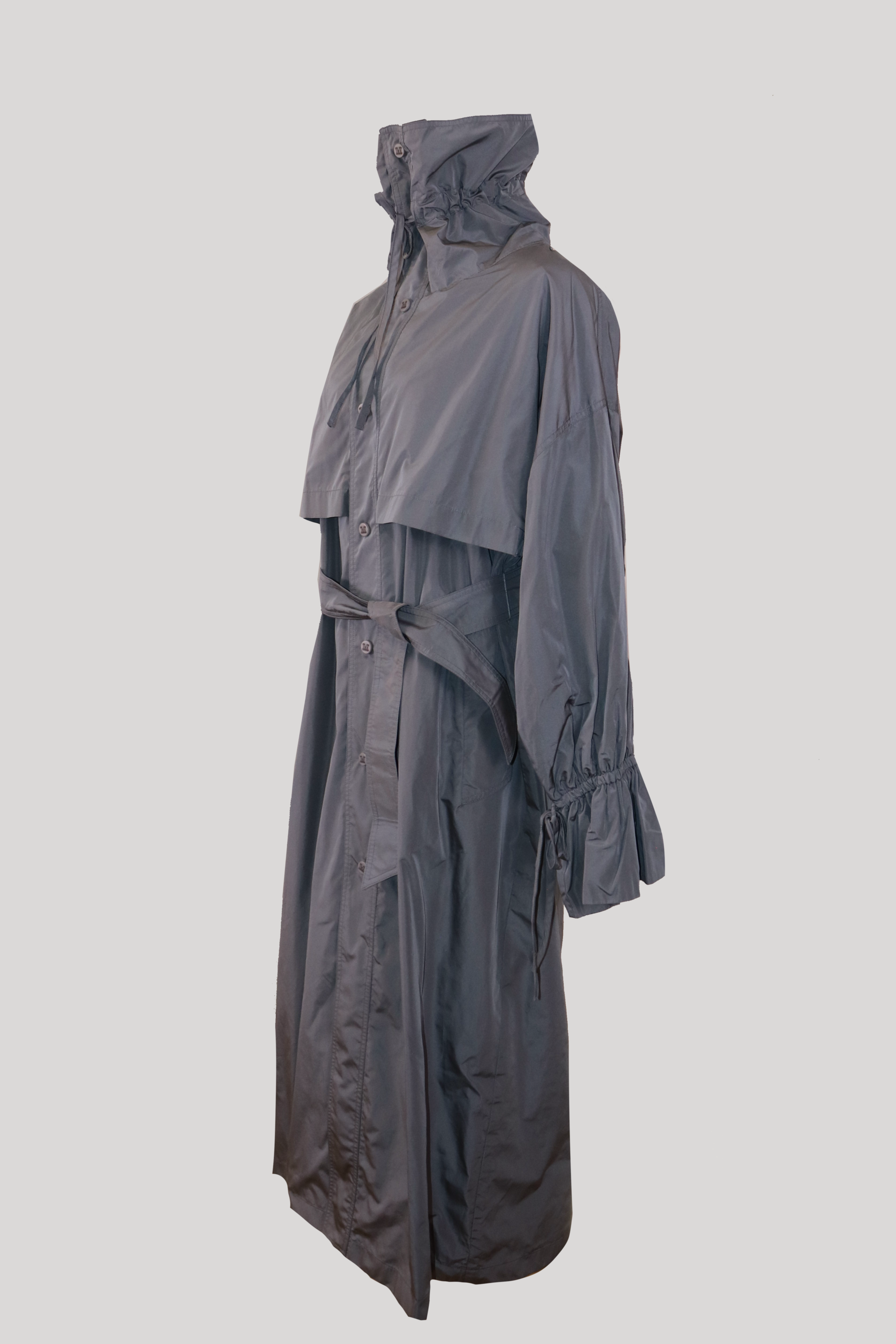 profile view of a grey high-neck max mara polyester trench coat. shows a relaxed silhouette, dropped shoulders, structural chest storm flaps, and loose sleeves with elasticized tie cuffs.