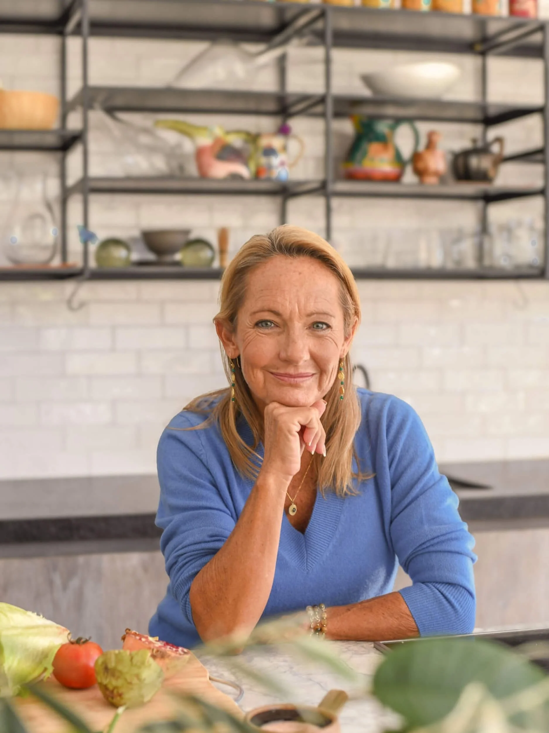 An older woman with blonde hair, wearing a blue sweater, smiling and resting her chin on her hand at a kitchen table. There are fruits and vegetables on the table and decorative shelves with colorful pots and dishes in the background.