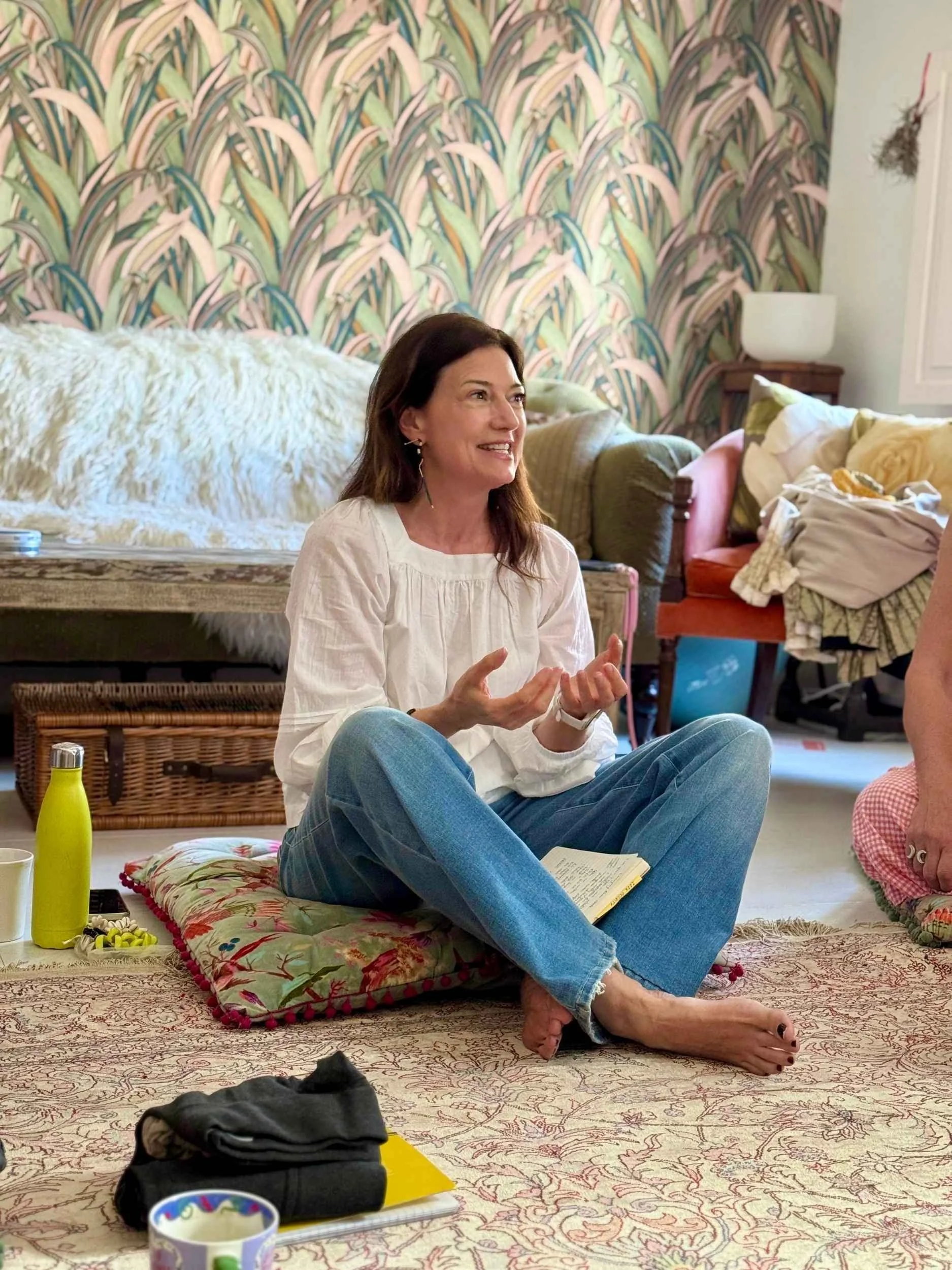 A woman with dark brown hair and earrings sitting cross-legged on a patterned rug, smiling and gesturing with her hands. She is wearing a white blouse and blue jeans. The room has a colorful, patterned wallpaper and various cushions and belongings scattered around, with a wooden table, a lamp, and a side table in the background.