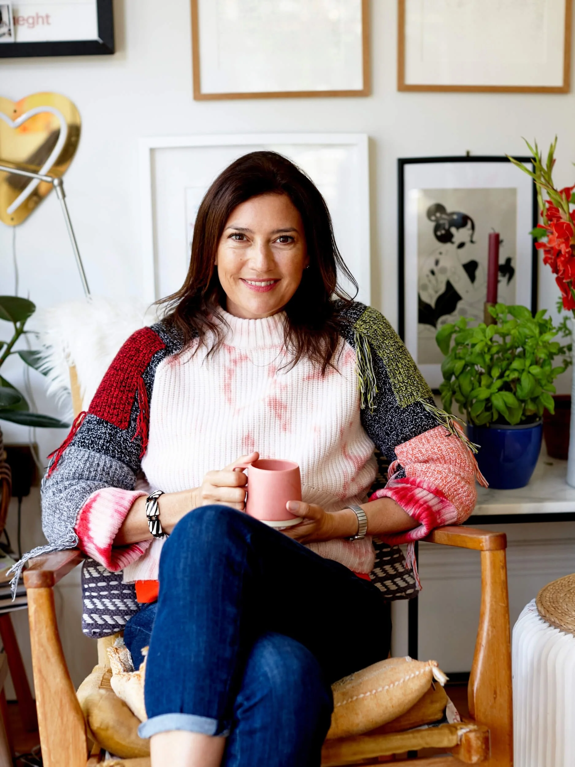 Woman sitting in a wooden chair, holding a pink mug, smiling at the camera, surrounded by plants and framed artwork on the wall.