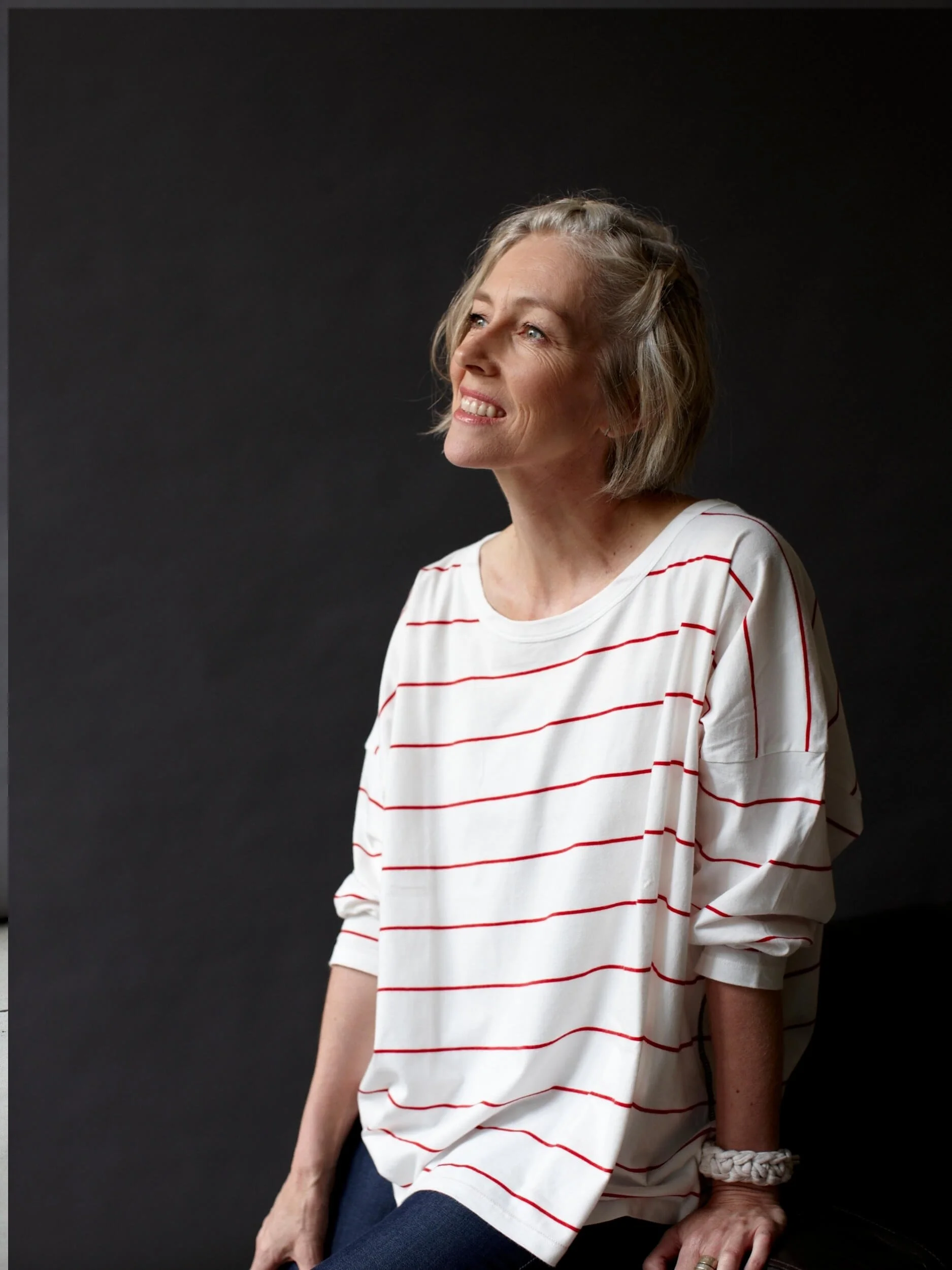 A smiling woman with short blonde hair, wearing a white shirt with red horizontal stripes, sitting against a black background.