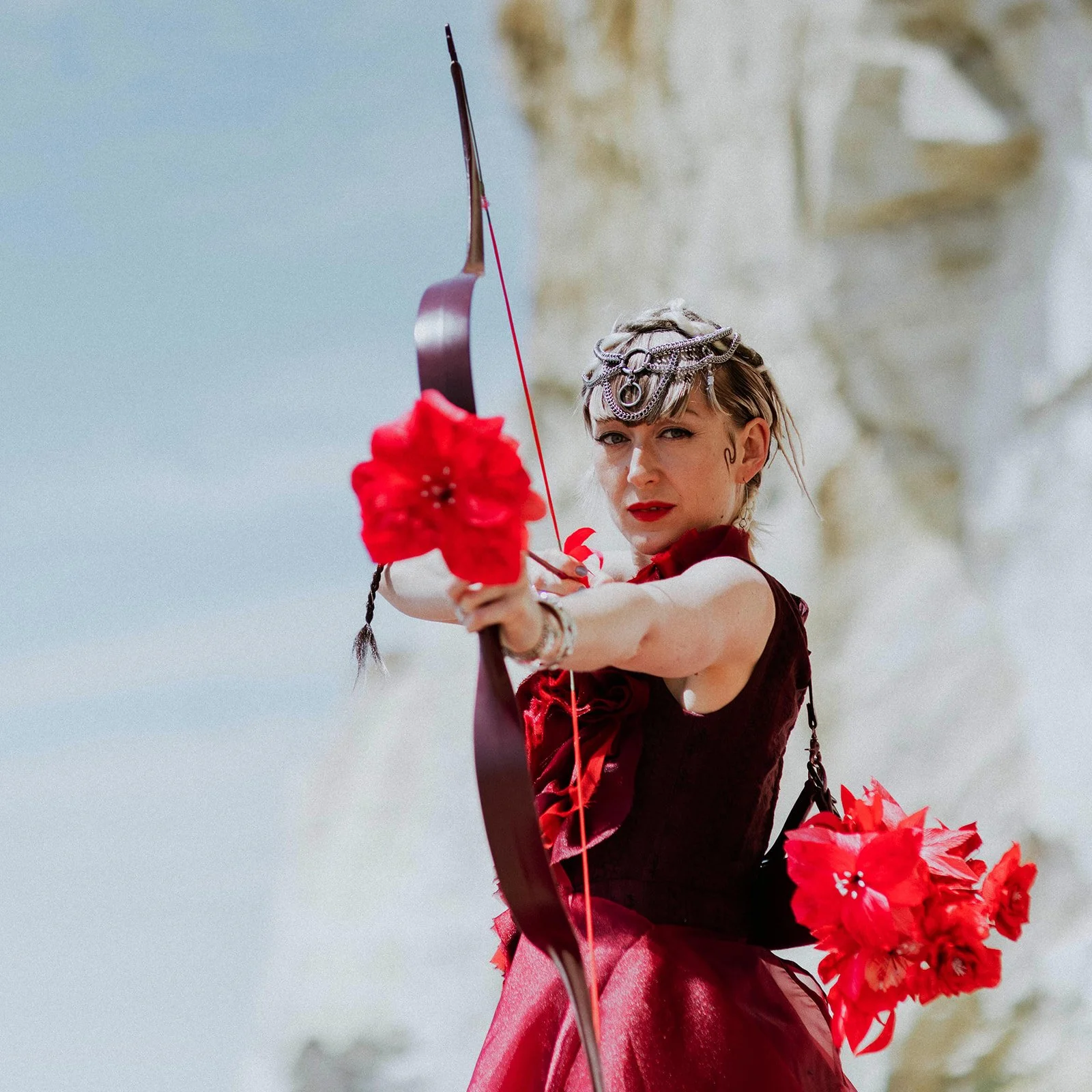 A person dressed in a red outfit with decorative headwear and holding a bow and arrow with a red flower attached, aiming outdoors.