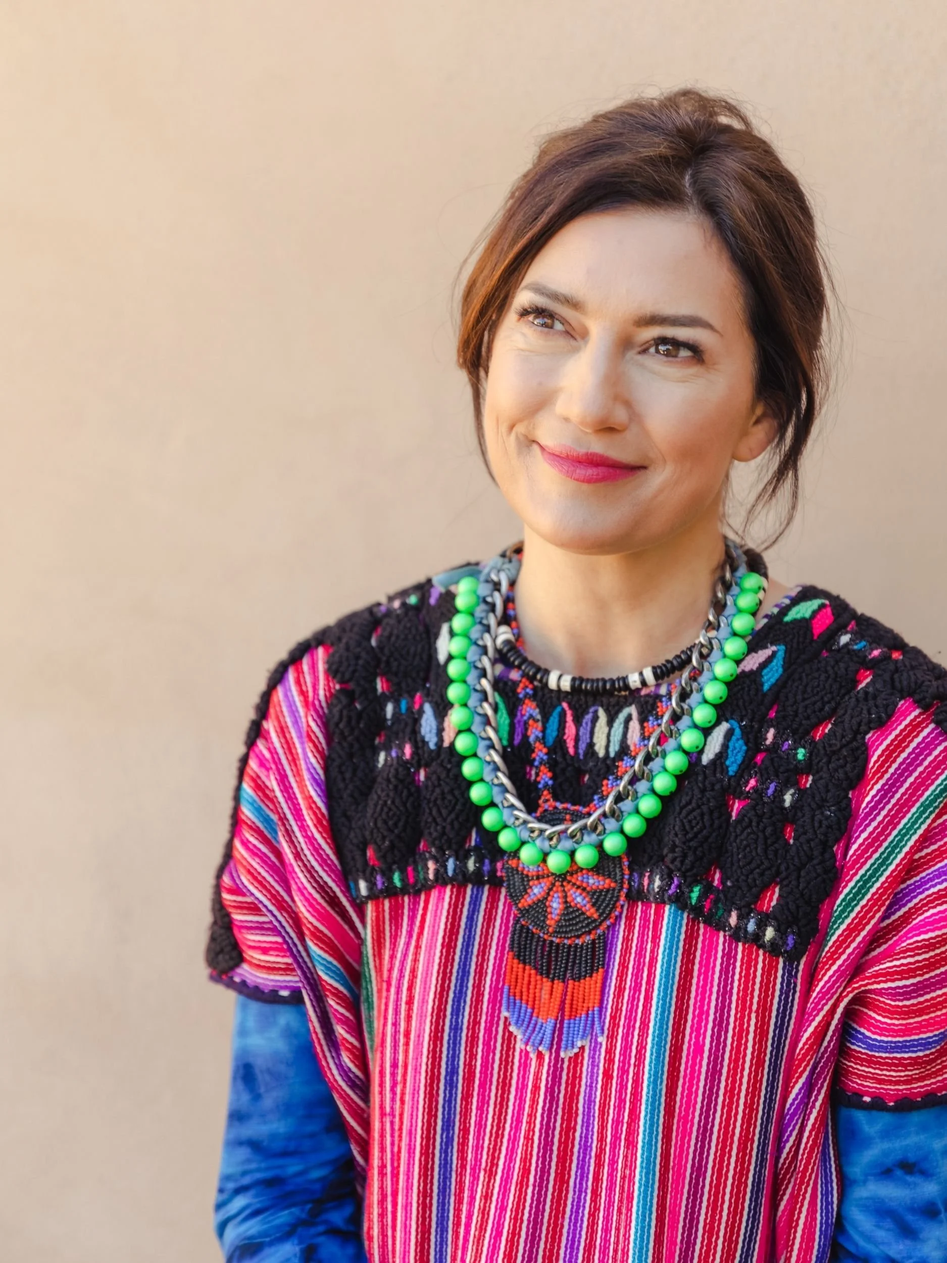 Woman wearing traditional Mexican colorful embroidered blouse with bright pink, red, green, purple, and blue stripes, accessorized with layered necklaces and a butterfly pendant, standing against a plain neutral background.