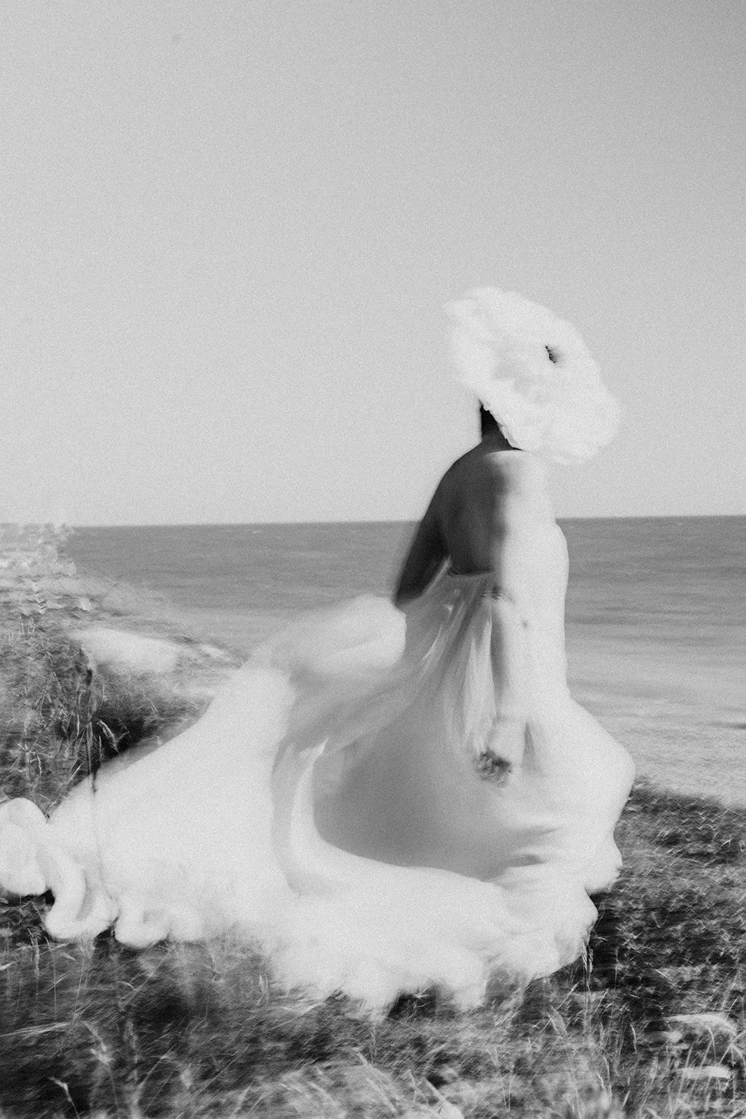A person with a large umbrella stands on a rocky shoreline by the ocean, wearing a flowing dress. The image is black and white.