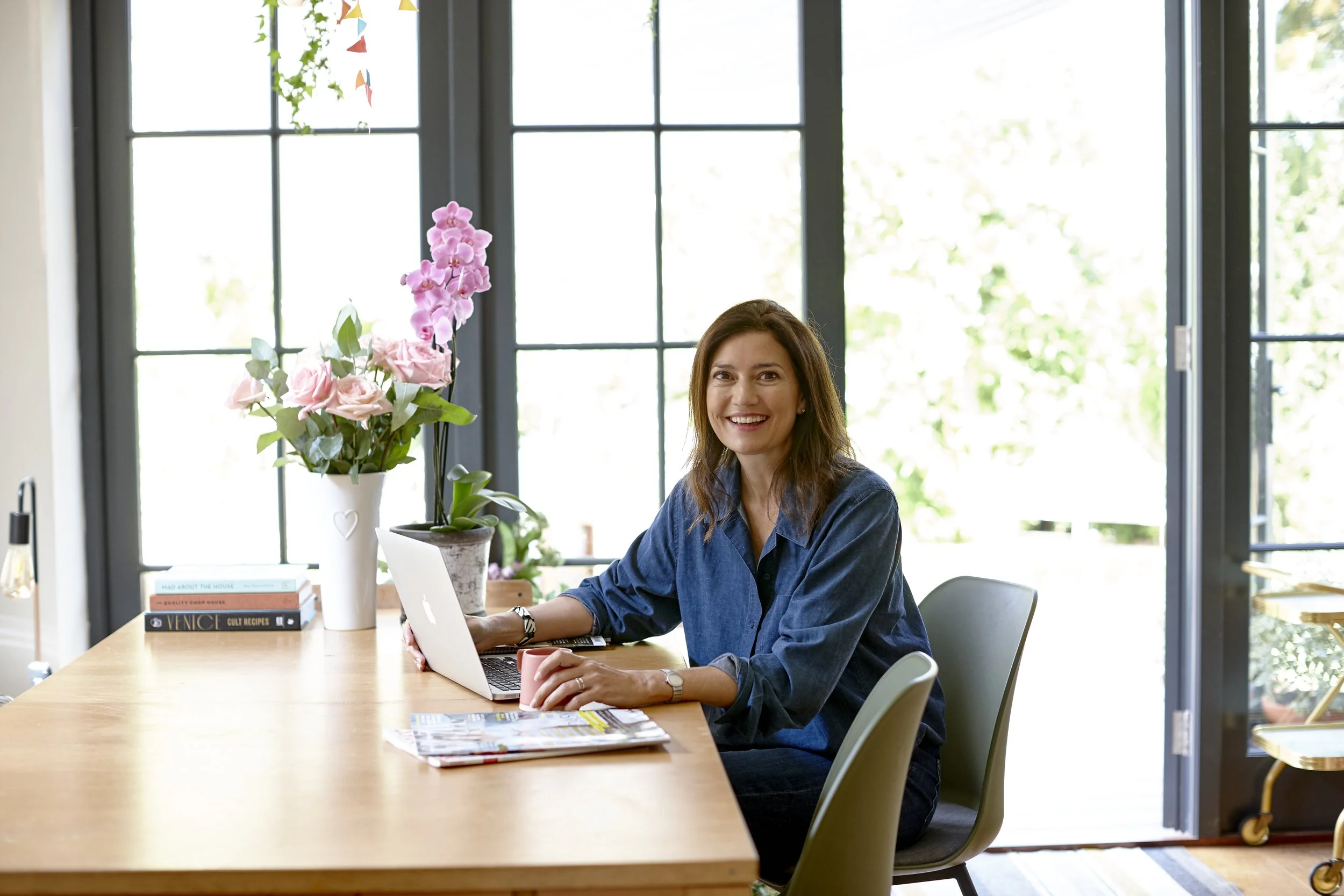 A woman with brown hair sitting at a dining table in front of large windows, smiling at the camera. There are pink flowers and books on the table, along with a laptop and a pink mug. The room is brightly lit with natural light.