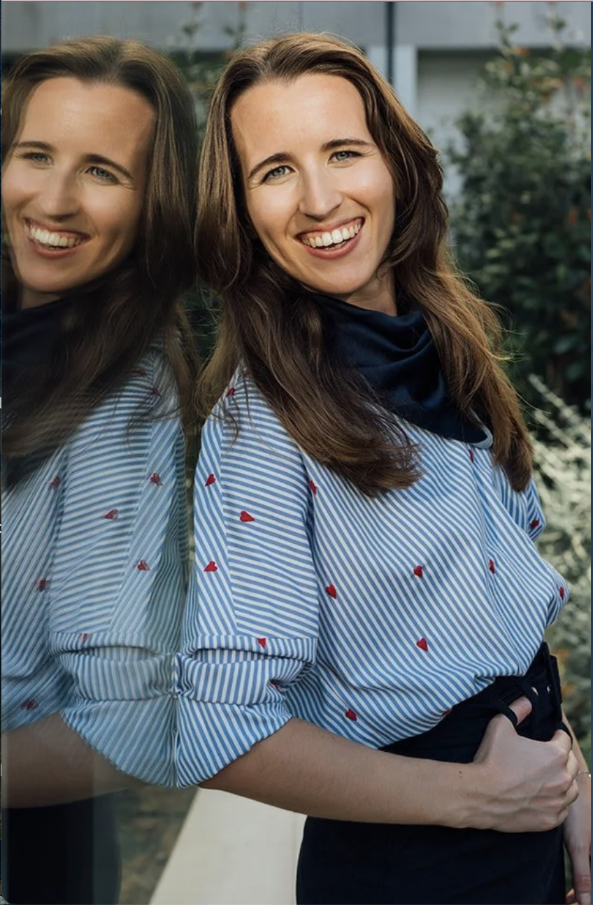 Two women standing side by side outdoors, one with dark brown hair smiling and wearing a blue and white striped shirt with small red hearts, the other with a reflection visible in a mirror, also smiling and wearing a similar shirt. The background includes greenery and a building.