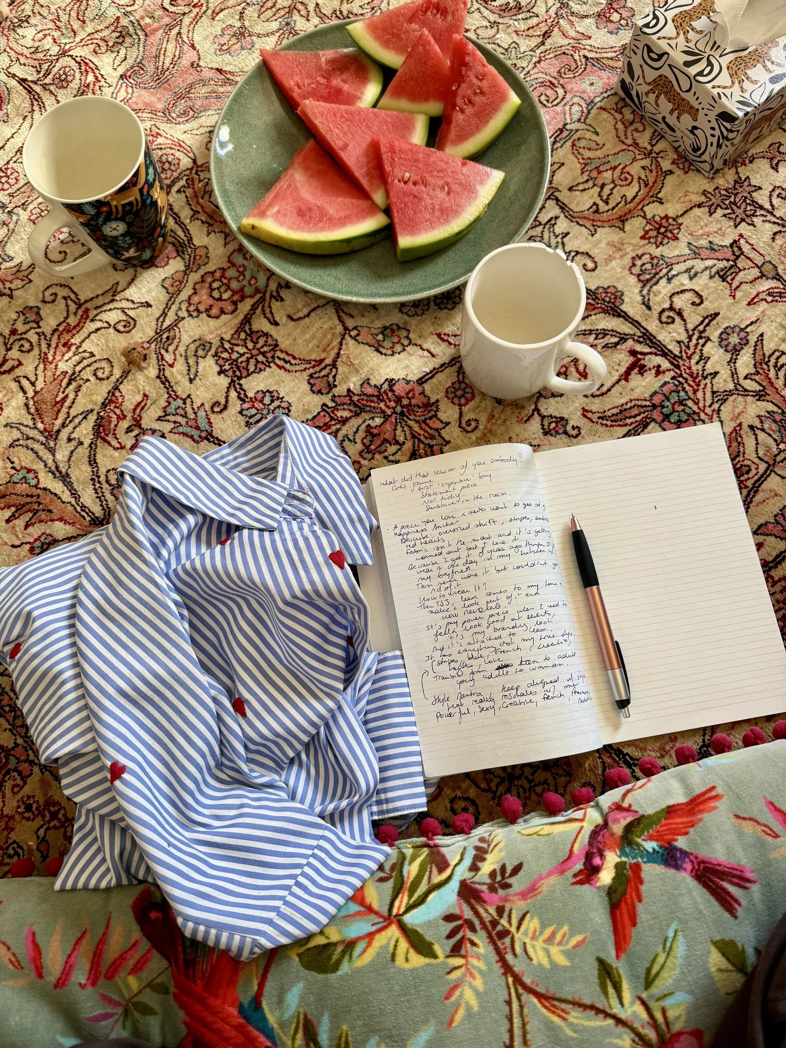 A tablecloth with a floral pattern in shades of red, pink, green, and beige, holds half a striped shirt with red heart accents, an open notebook with handwritten notes, a black and rose gold pen, two empty coffee mugs, a green plate with watermelon slices, and a tissue box with animal prints.