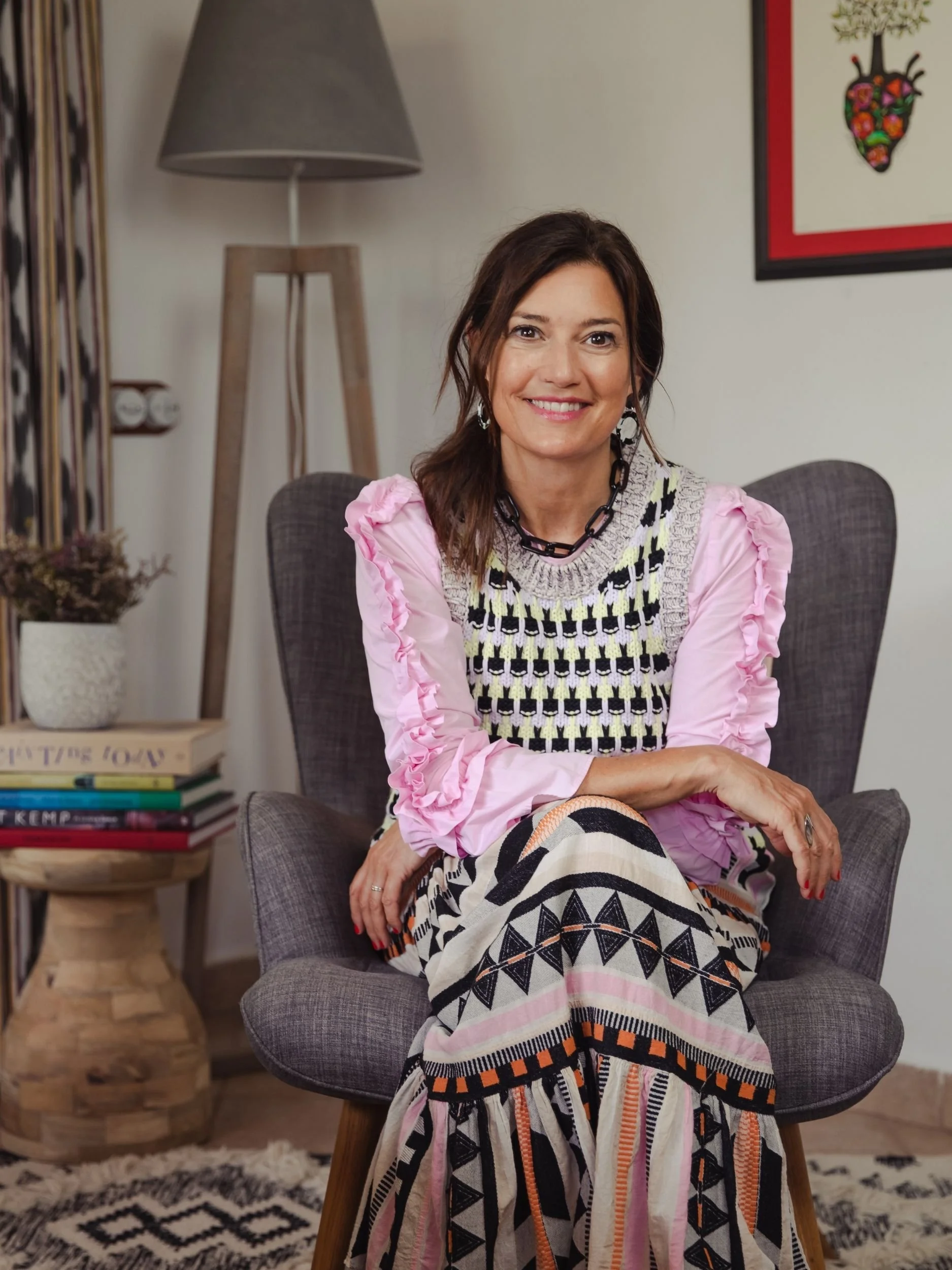 A woman with dark brown hair and a warm smile, wearing a patterned dress with puffy pink sleeves, sitting in a modern gray armchair in a cozy living room. Behind her is a framed colorful art piece featuring a heart-shaped design with floral elements, a tall floor lamp, a side table with a stack of books, and a potted plant.