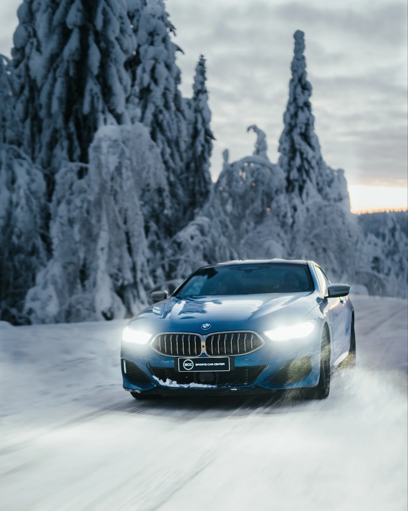 BMW m8 auto ajaa lumisella tiellä metsän keskellä tykkypuiden ympäröimänä.