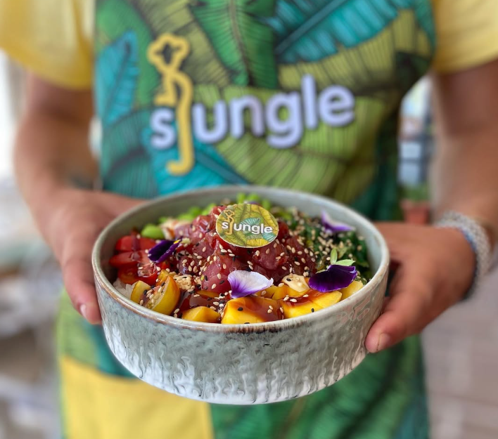 Una persona, indossando una maglietta con il logo 'Sjungle', tiene in mano una ciotola con un piatto di poke. Il piatto è valorizzato e facilmente identificato dalla cialda alimentare posta al centro che riporta il logo del brand