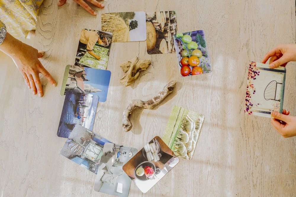 People playing a card game with landscape and still life photos on a wooden table, with two hands visible holding cards and some pieces of driftwood on the table.