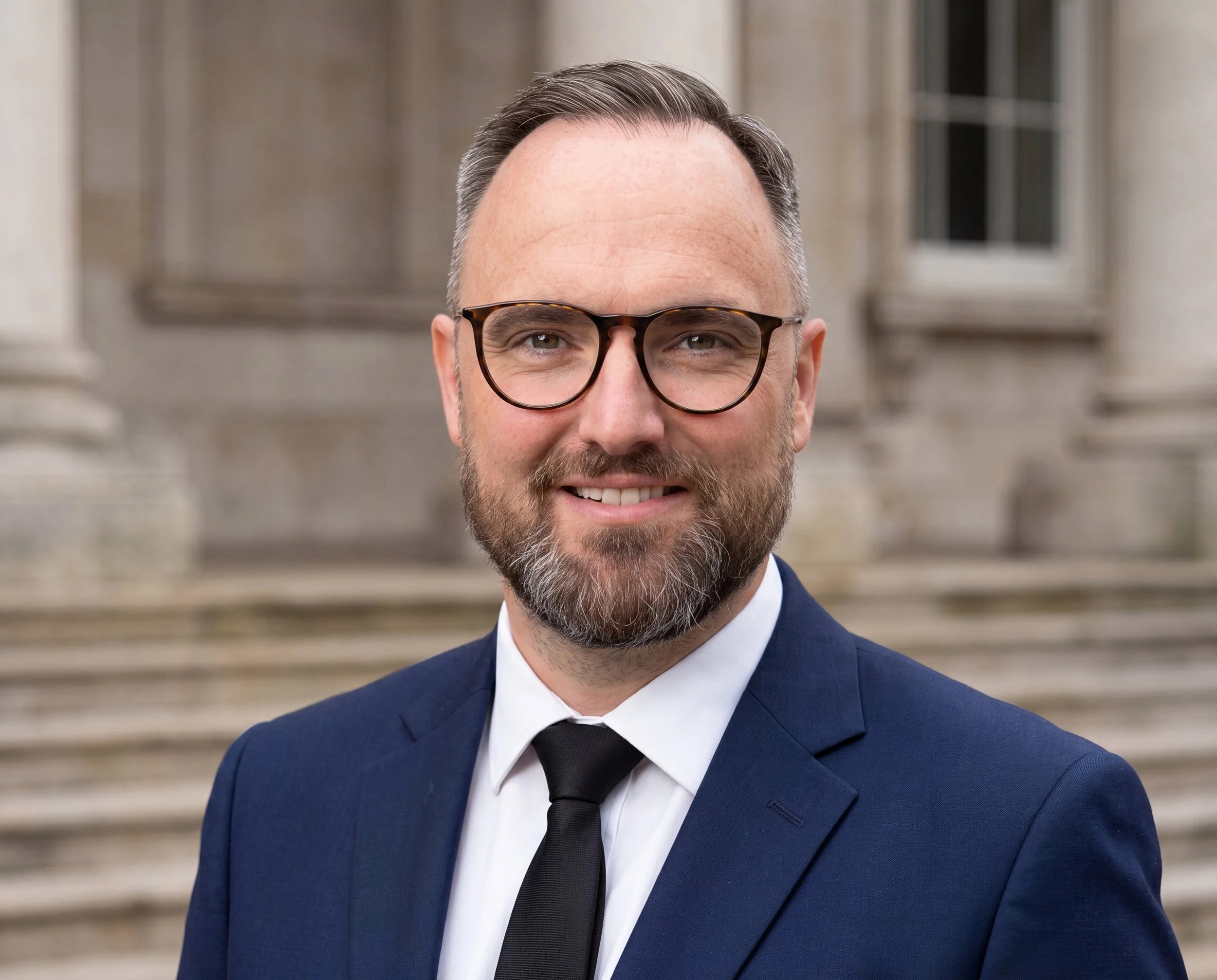 Close-up of a man with glasses, a beard, wearing a dark blue suit, white shirt, and black tie, smiling outdoors with a stone building in the background.