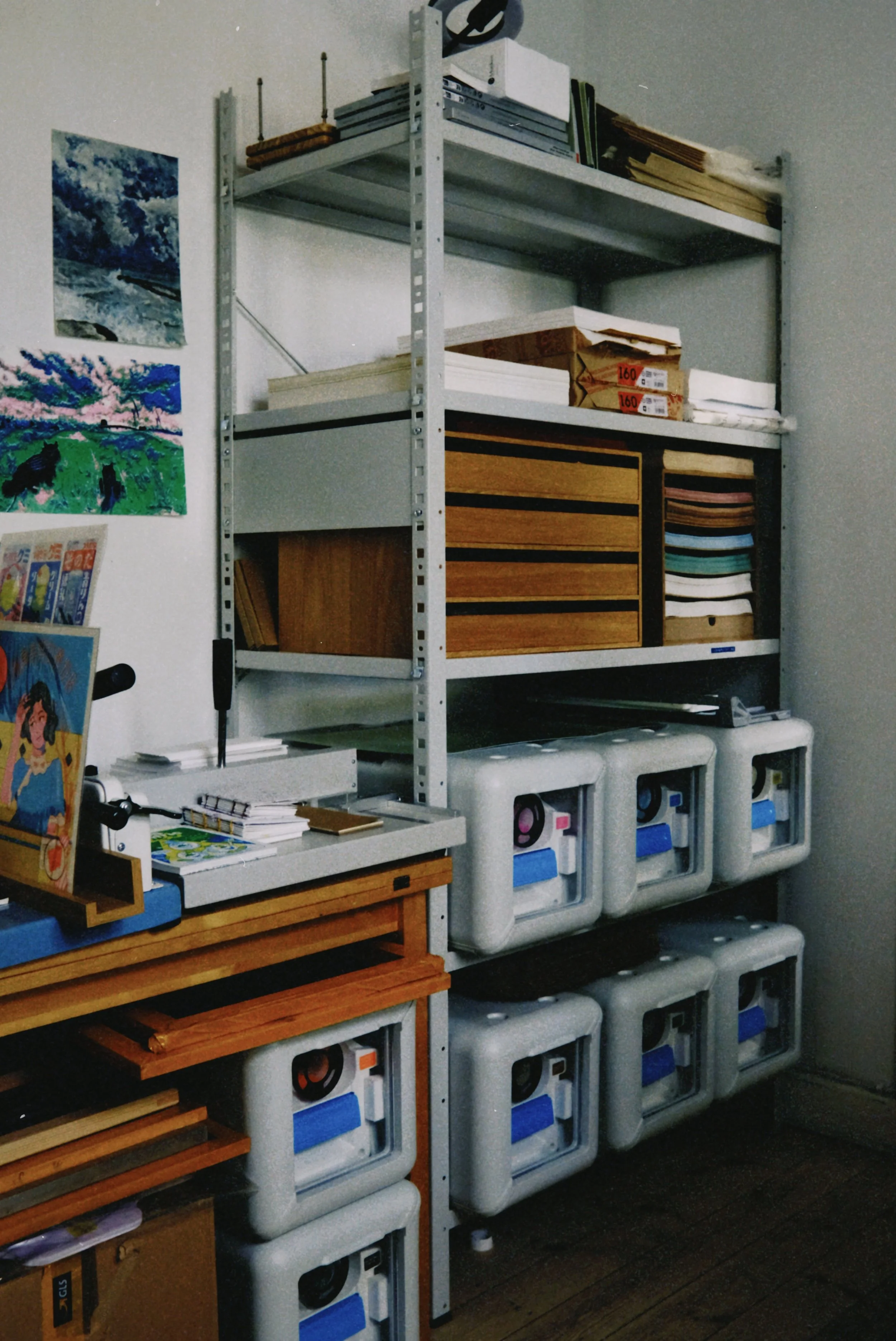 A metal shelving unit with art supplies including paper, magazines, and boxes. On the wall are watercolor paintings of a waterfall and a tree with pink blossoms. There are also cartoons and books on a small wooden table in front of the shelving unit.