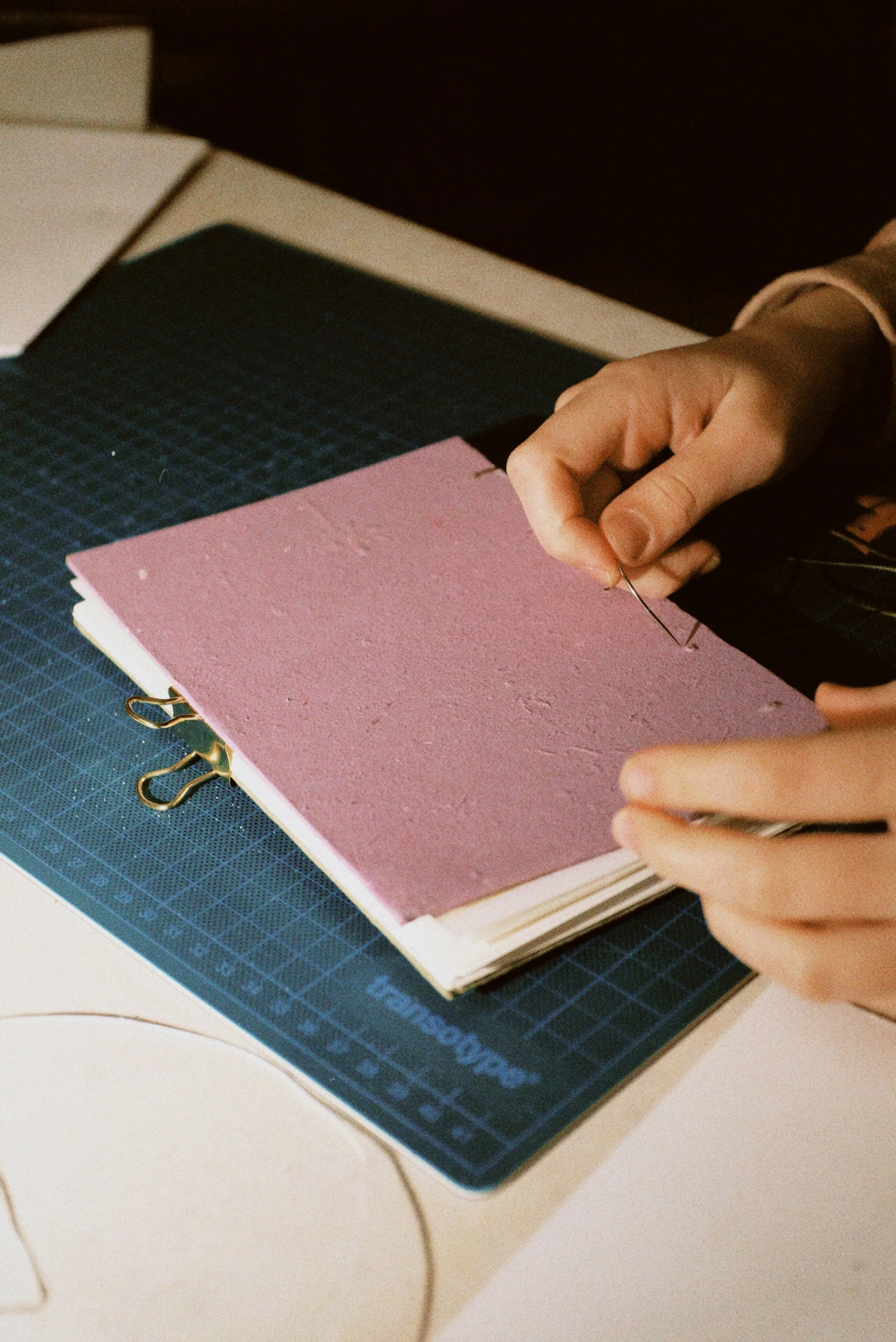 Person holding a needle and pink paper or fabric over a notebook, on a work table.