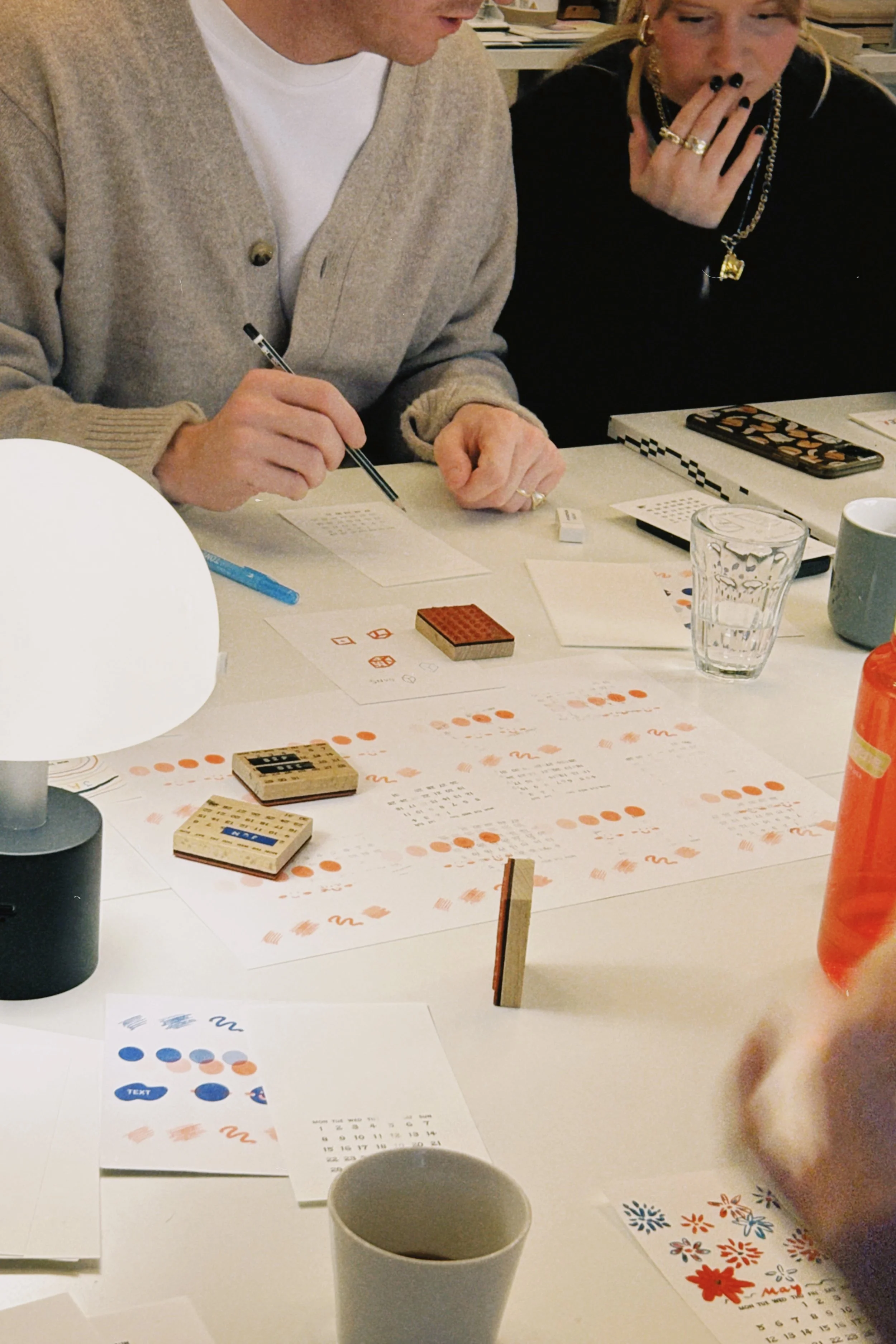 People engaging in a tabletop game with bingo cards, markers, and various small game pieces, surrounded by drinks and assorted stationery.
