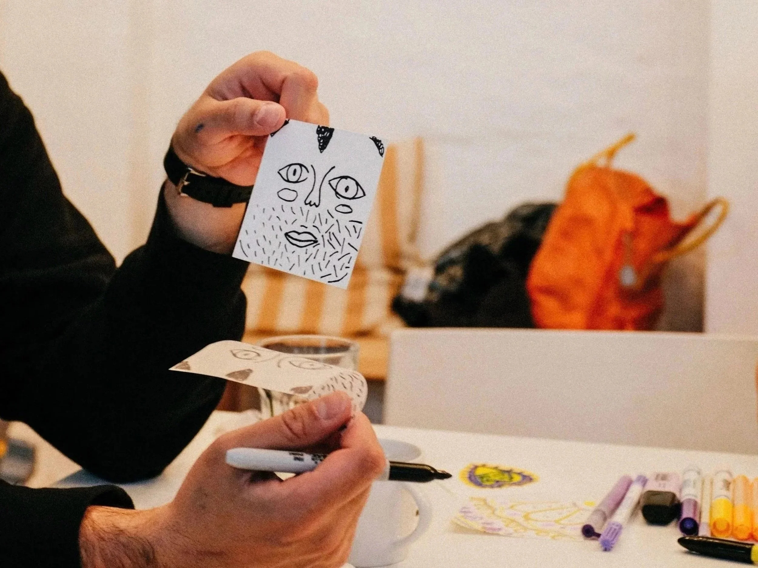Person holding a small piece of paper with a line drawing of a face, at a table with colored markers and other drawings.