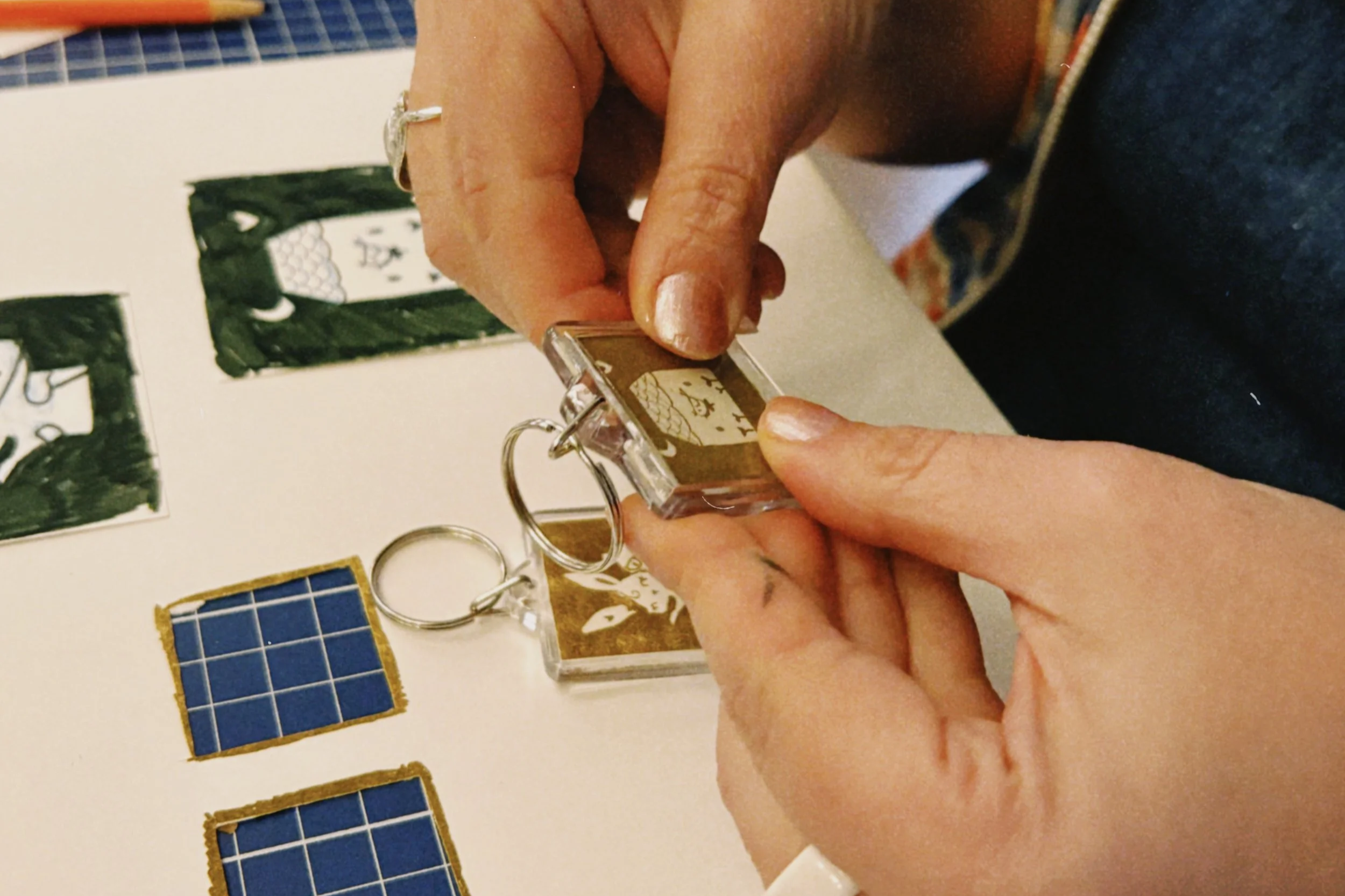 Person holding a keychain with a clear plastic casing containing a design, near some small artwork pieces on a white surface.