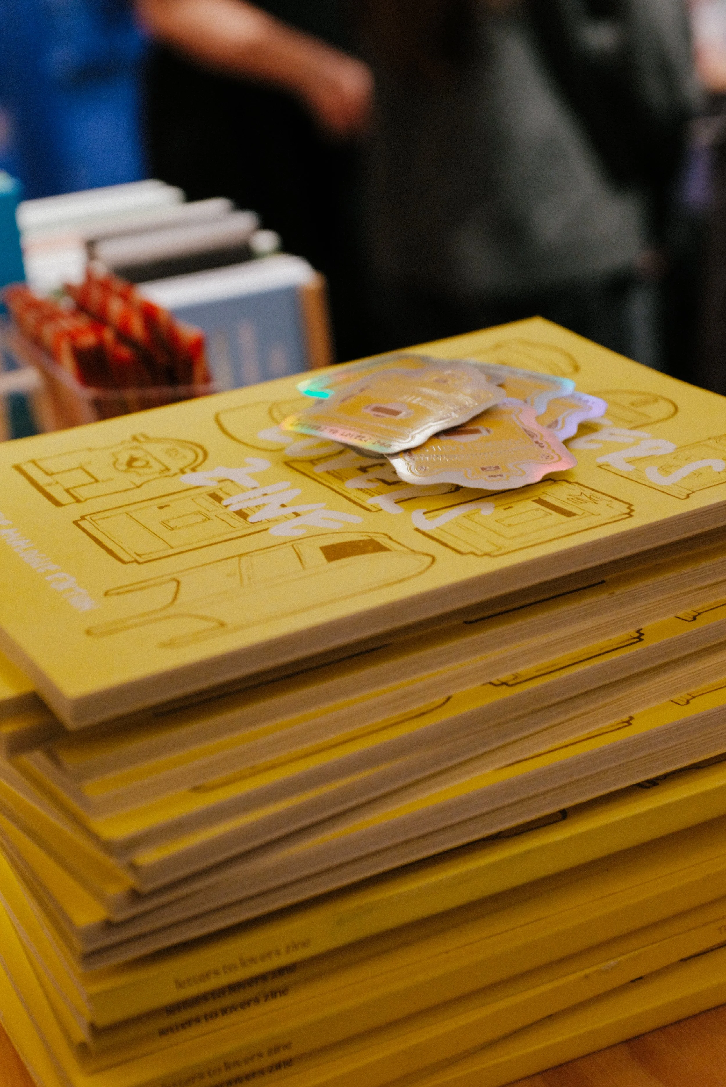 Stack of yellow brochures or pamphlets with small items on top, possibly stickers or small paper packets, in a setting that appears to be at a booth or display stand.
