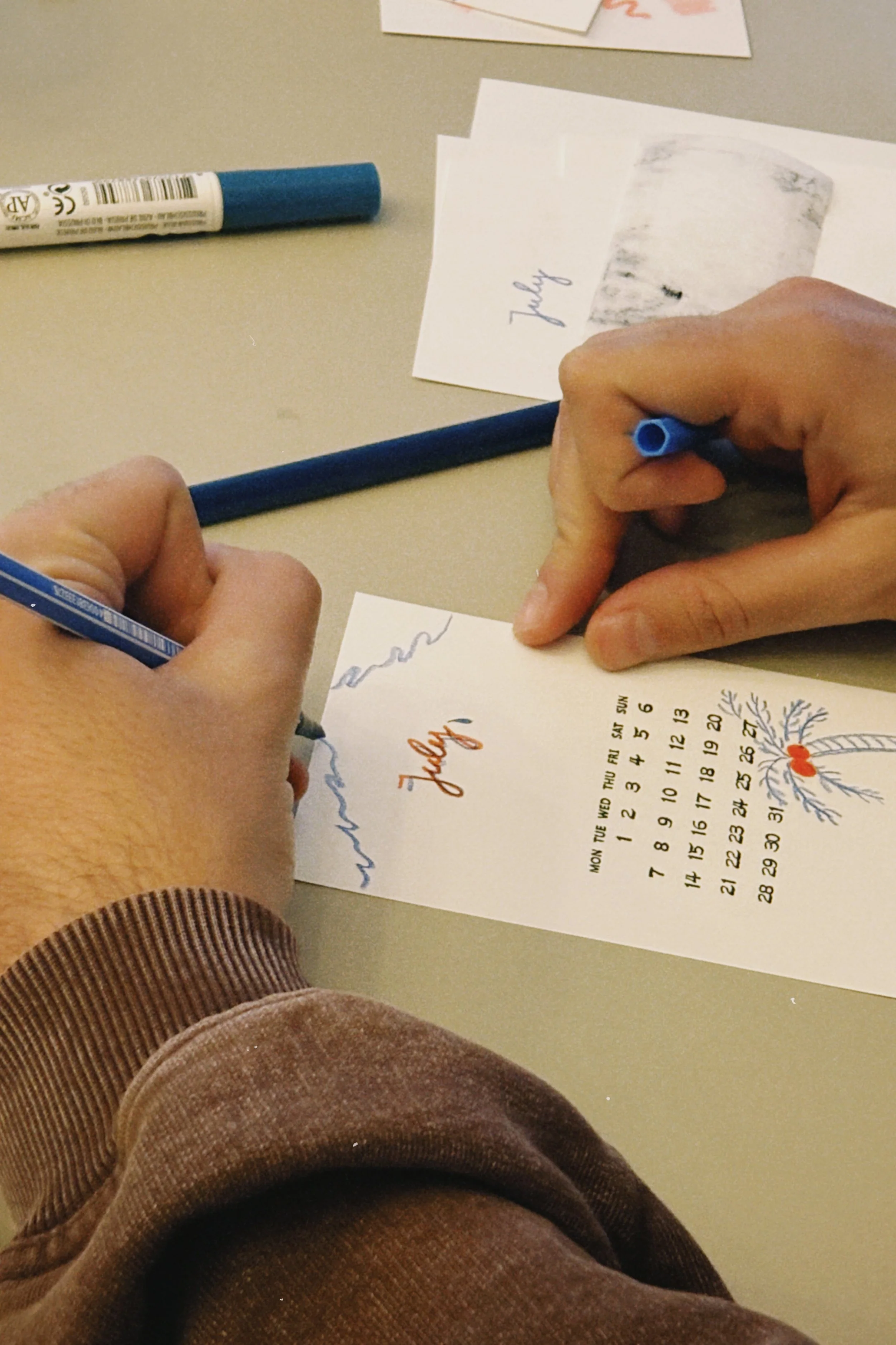 A person wearing a brown striped shirt writing on a piece of paper with a blue pen, a calendar with decorative elements on the page, and an additional note or card on the table.
