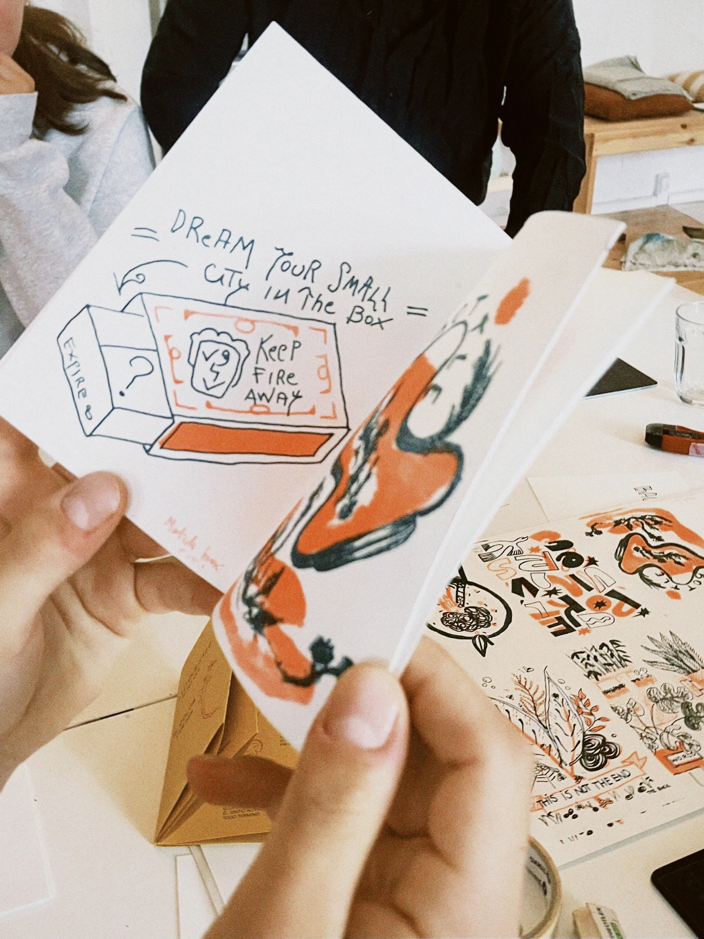 Close-up of a person holding a colorful illustrated card with hand-drawn text and images, with other cards and objects on a table in the background.