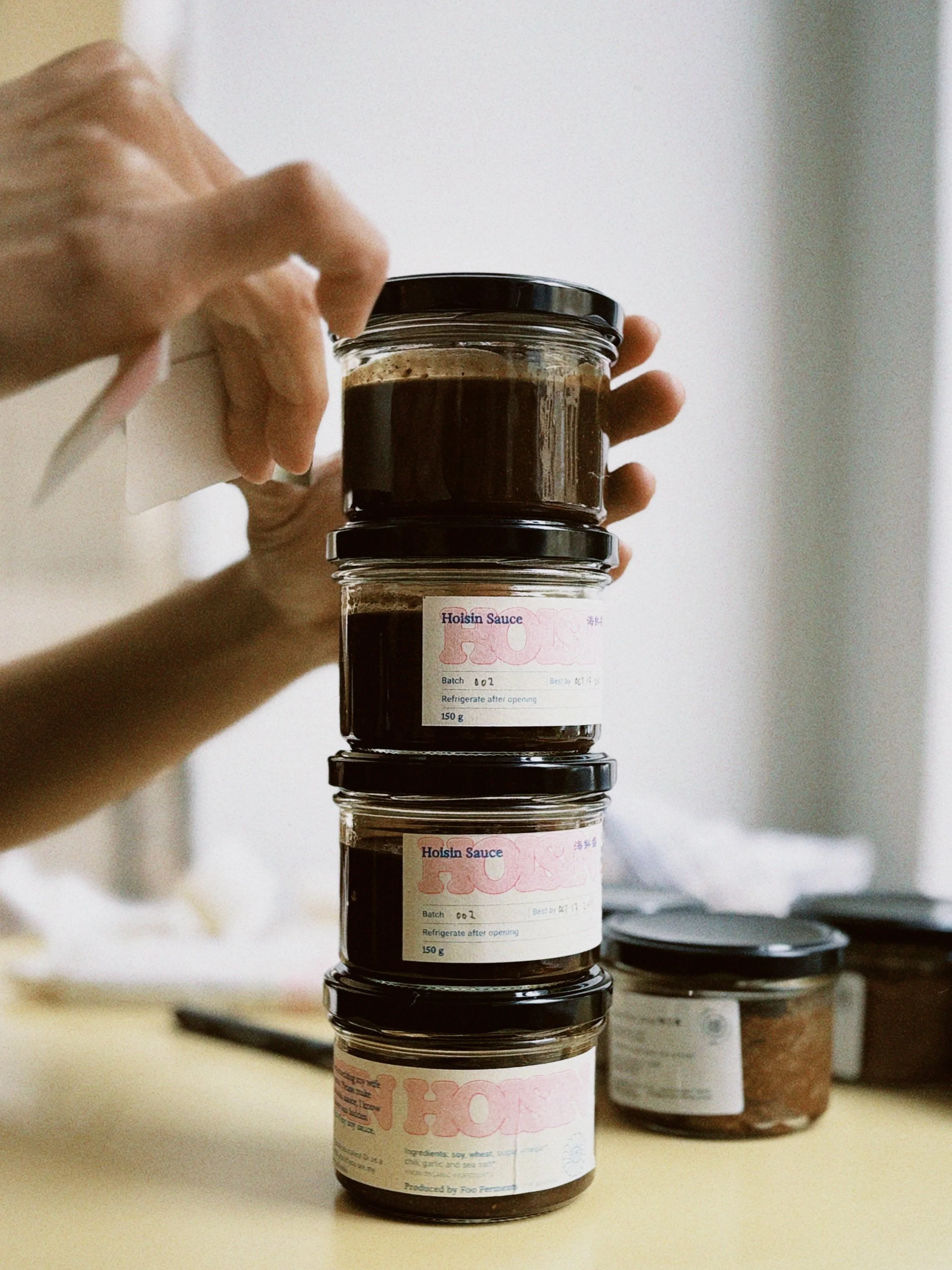 Stack of glass jars with black lids labeled Hoisin Sauce, placed on a light-colored surface with other jars in the background.