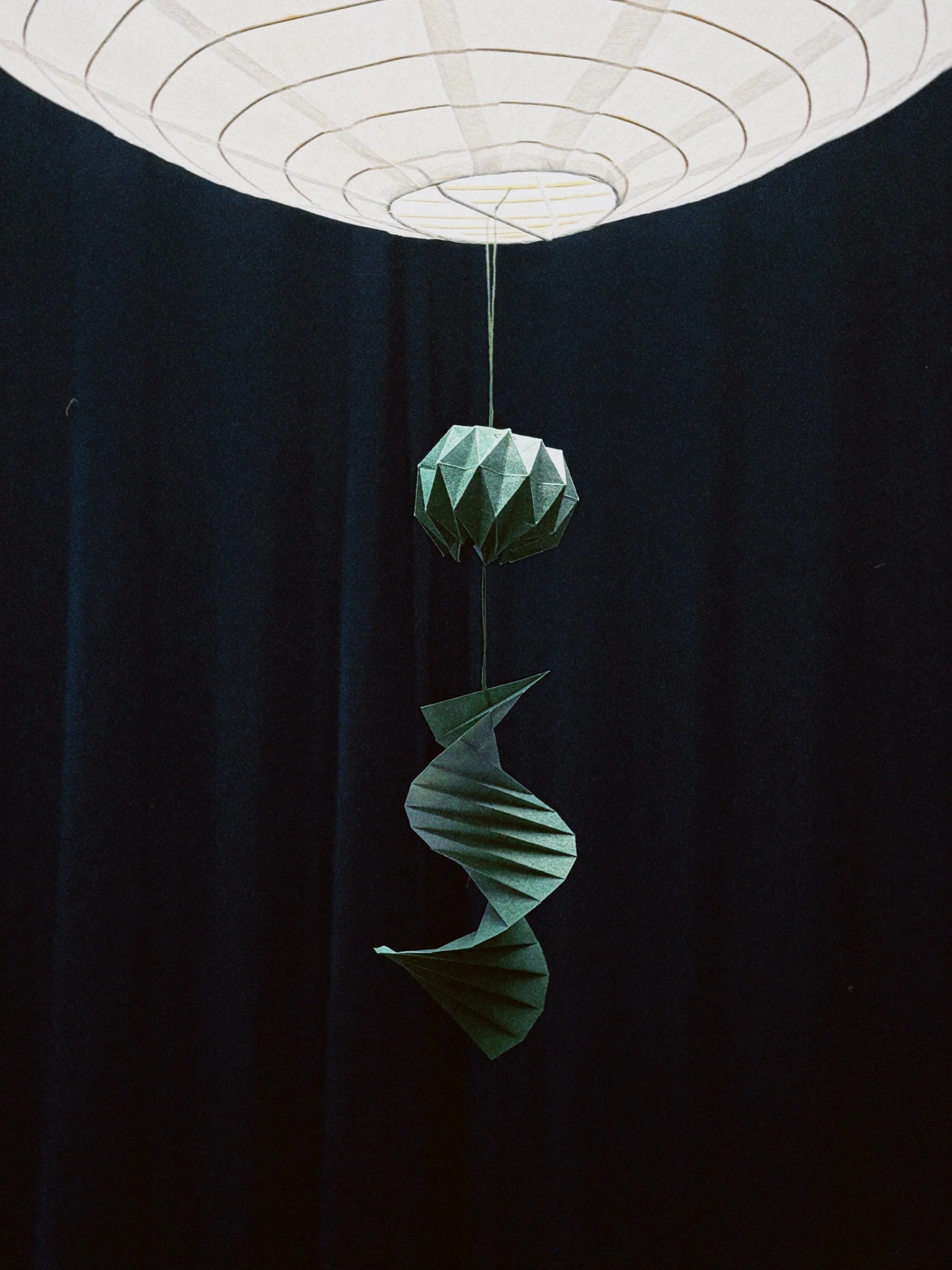 Hanging paper spiral decoration with a lotus flower shape, suspended from a white paper lantern against a dark background.