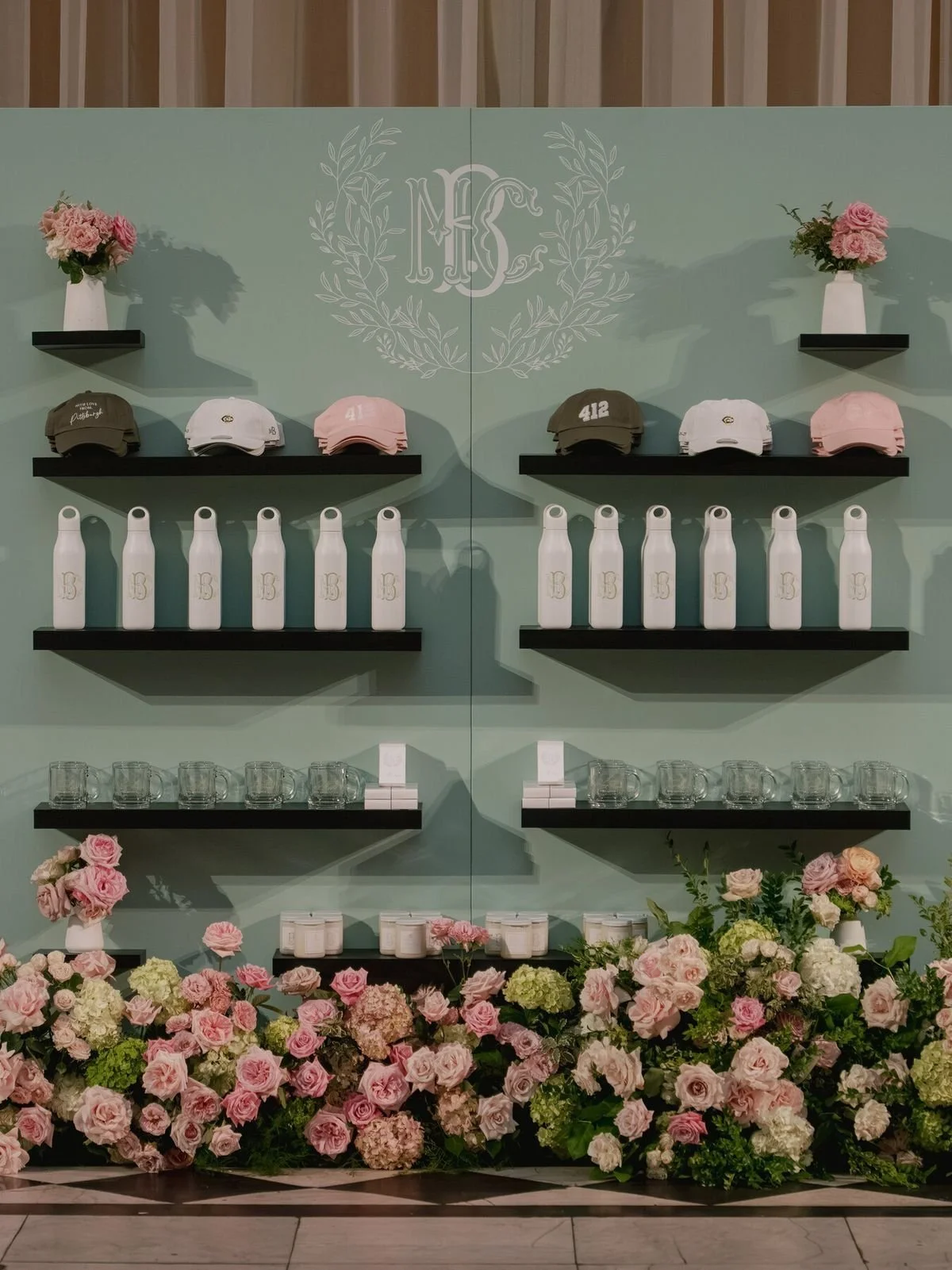 Display of pink and white flowers in vases, white caps with initials, black shelving with white water bottles, glass mugs, and candles against a mint green wall with a monogram logo.