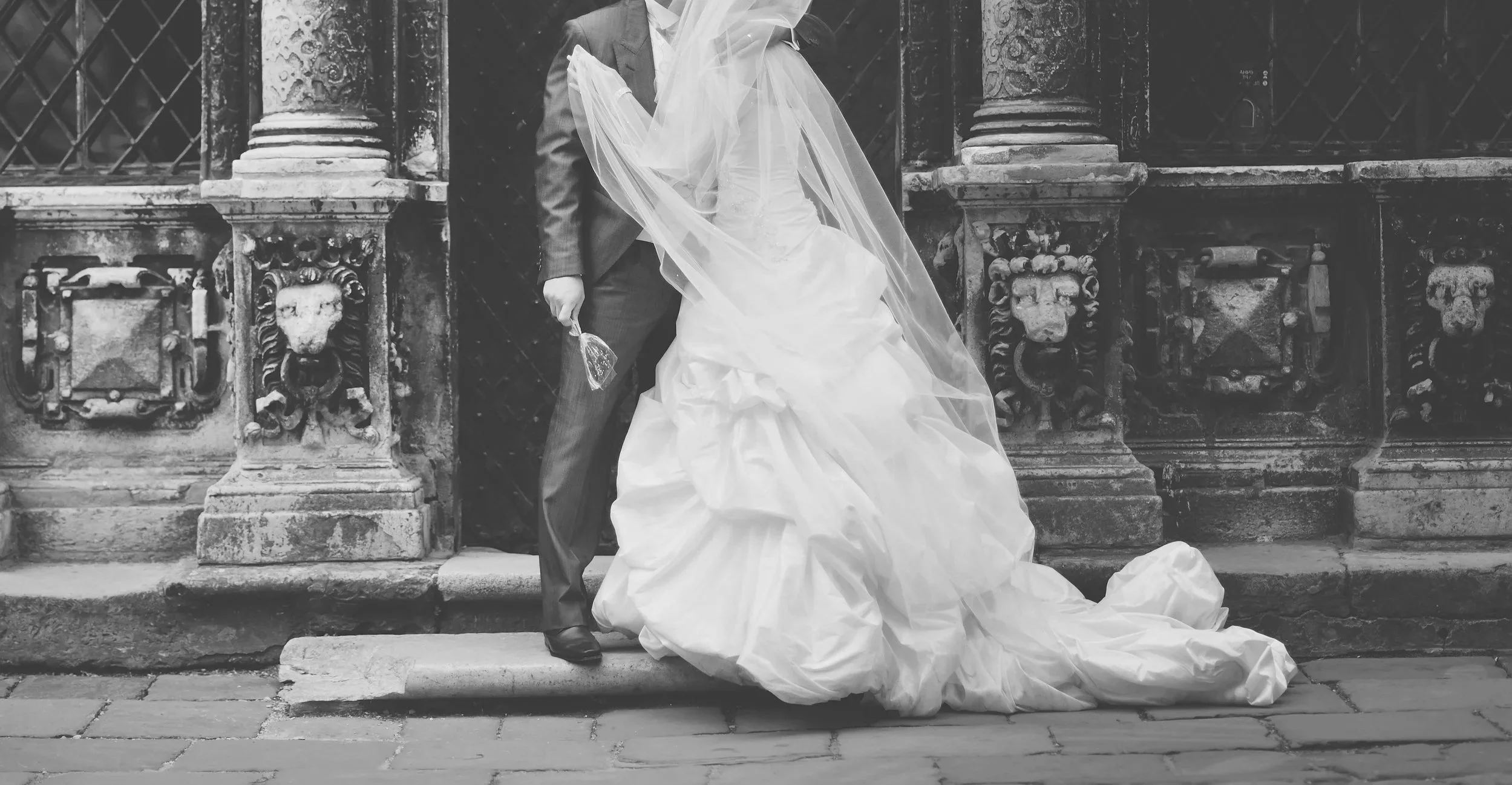 A black-and-white photo of a bride and groom standing close together on stone steps in front of an ornate, historical fence with columns. The bride is wearing a traditional white wedding dress with a long train and veil, and the groom is dressed in a suit, holding a small bouquet. Their faces are not visible in the image.
