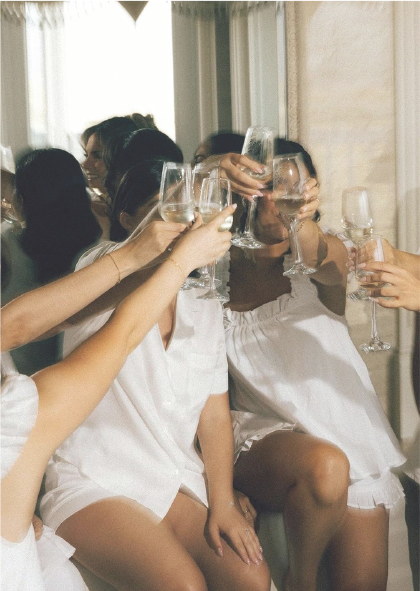 Group of women in white dresses raising wine glasses for a toast at a celebration.