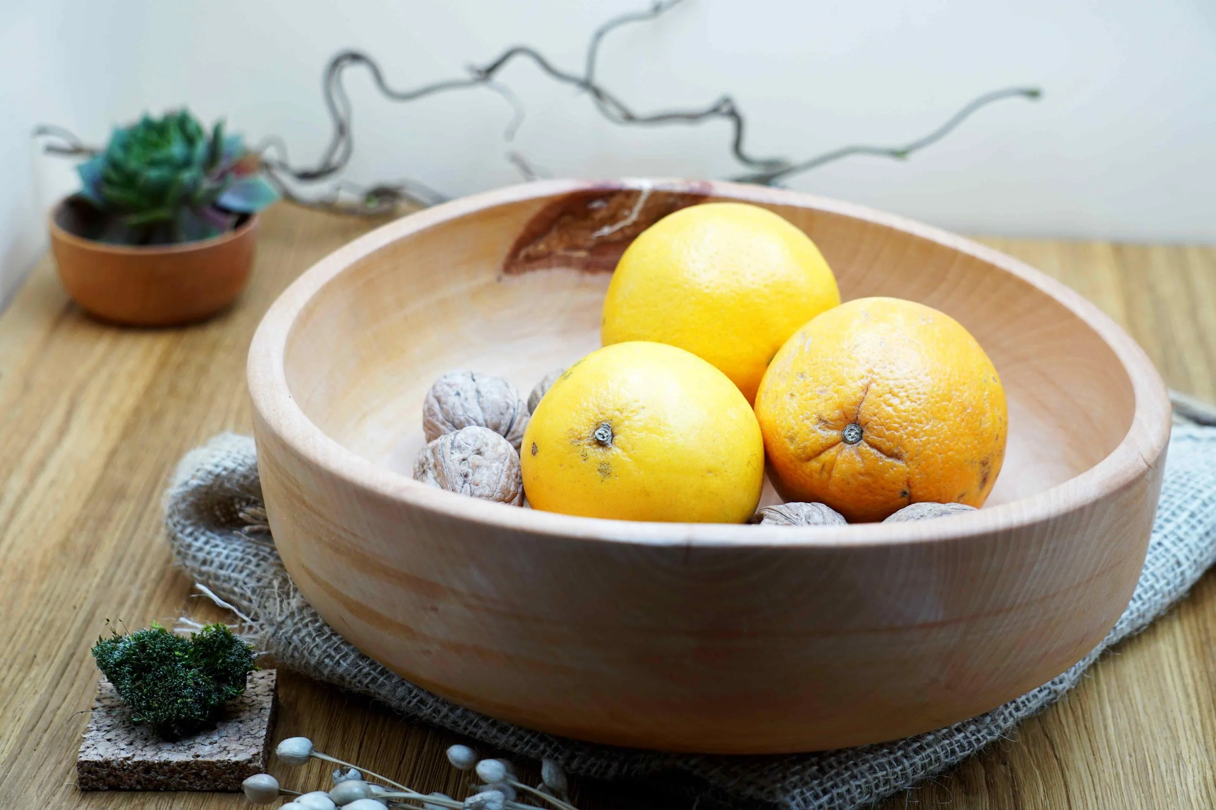 Runde Holzschale mit heller Oberfläche, gefüllt mit Zitrusfrüchten und Nüssen, platziert auf Holztisch mit textiler Unterlage.
