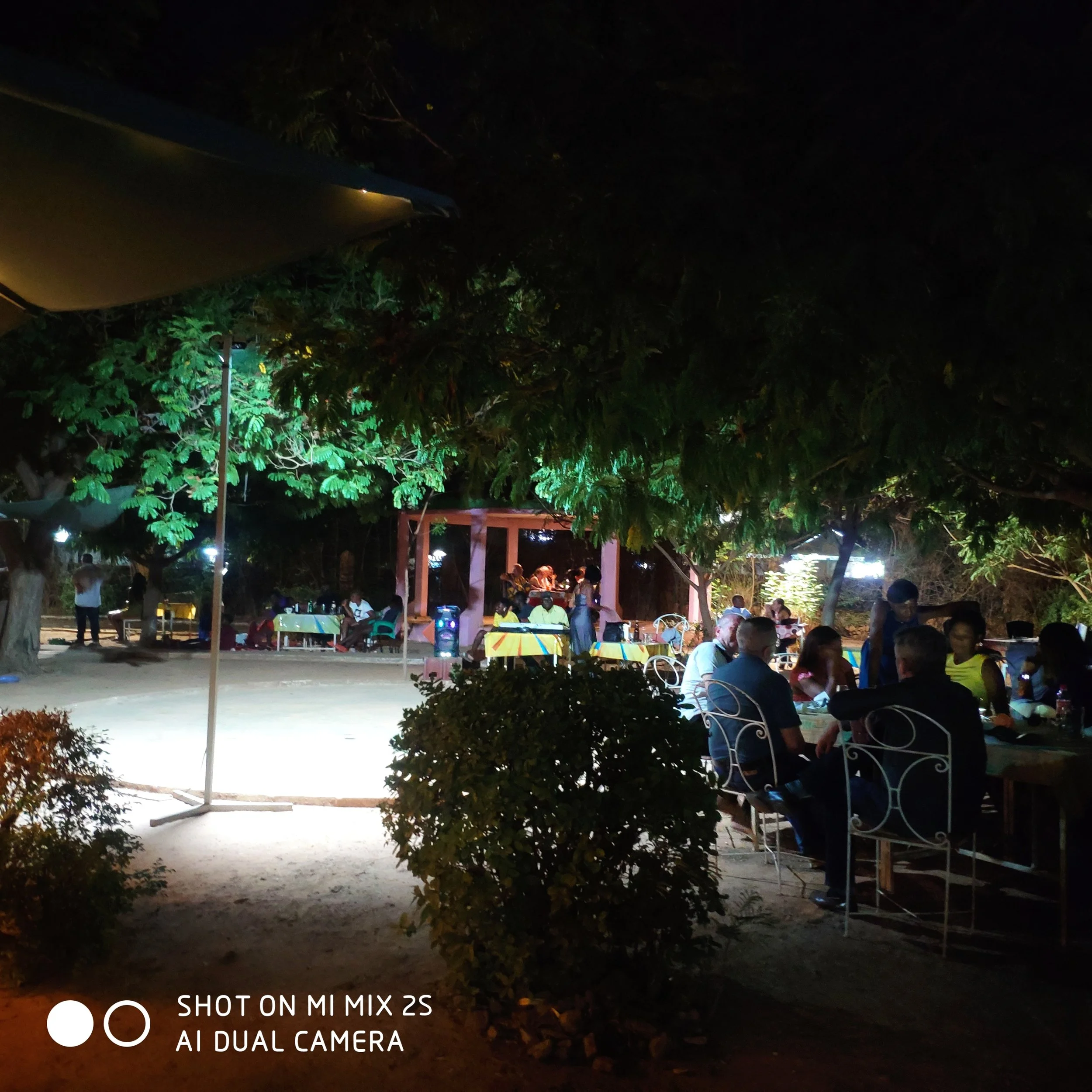 Une scène nocturne dans un jardin ou un parc avec des gens assis et discutant sous un abri en bois, entouré de plusieurs arbres et végétation, éclairée par des lumières artificielles.
