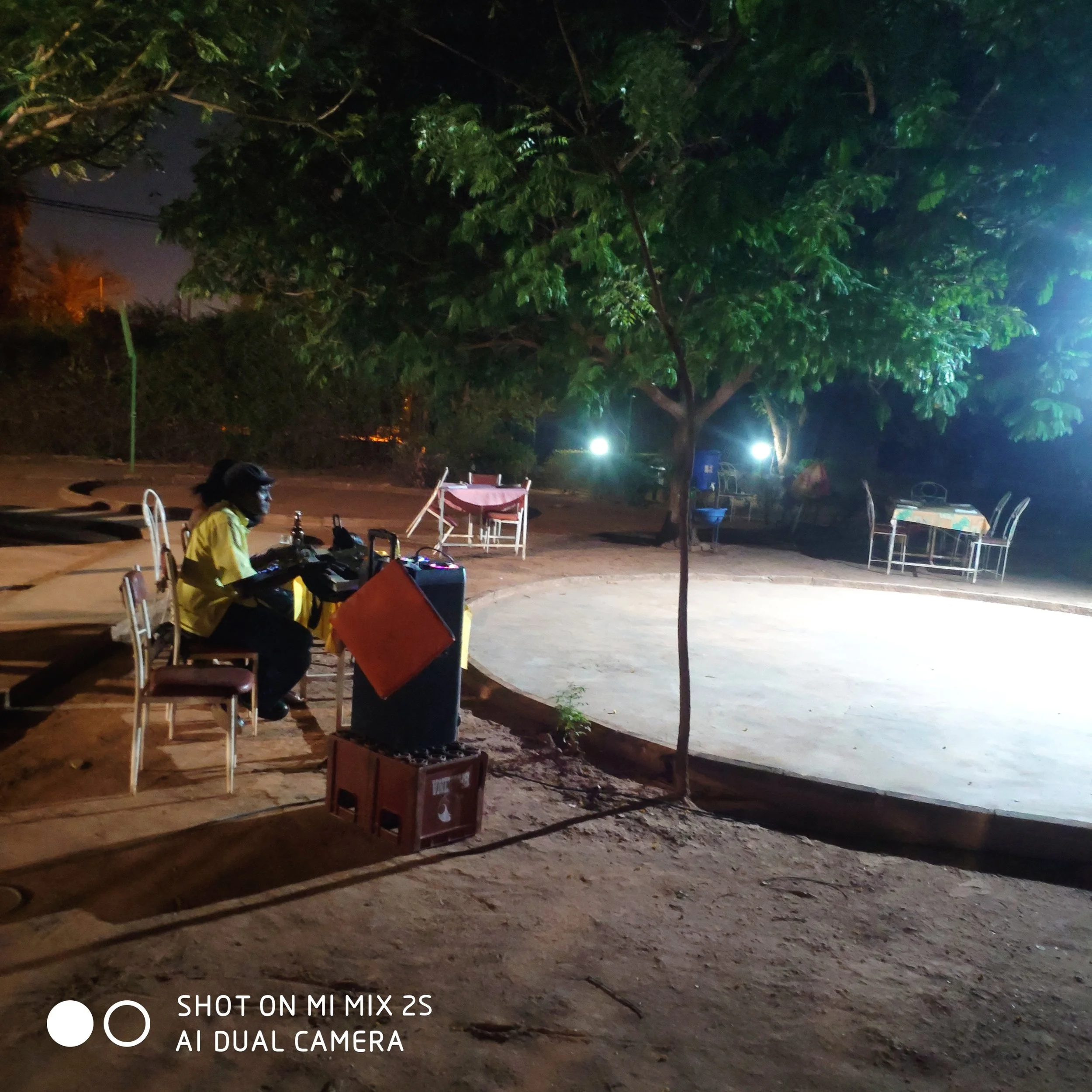 Vue nocturne d'une scène extérieure avec un grand arbre au centre, des lampadaires éclairant partiellement la scène, des tables et chaises, et une personne assise à une table. La photo a été prise avec un smartphone Xiaomi Mi Mix 2S, mode AI dual camera.