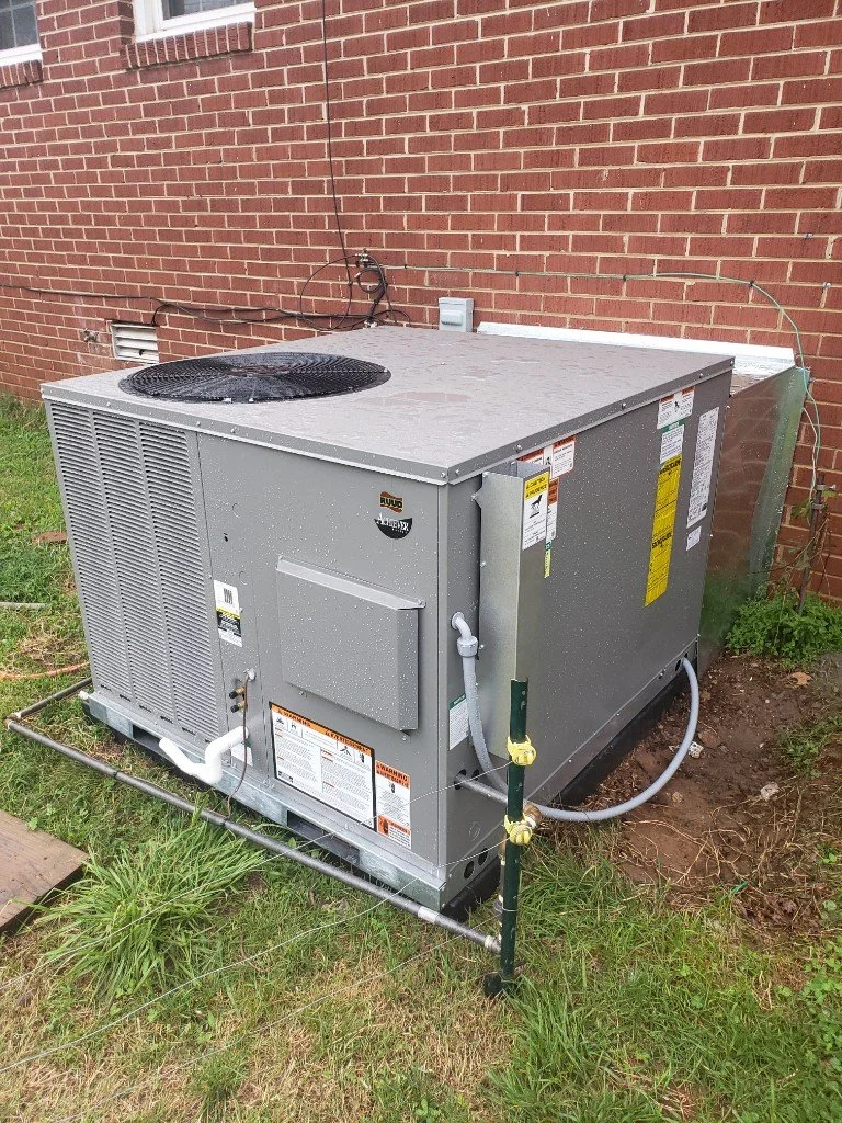 An outdoor air conditioning unit next to a brick wall, with pipes and electrical connections, installed on a patch of grass.