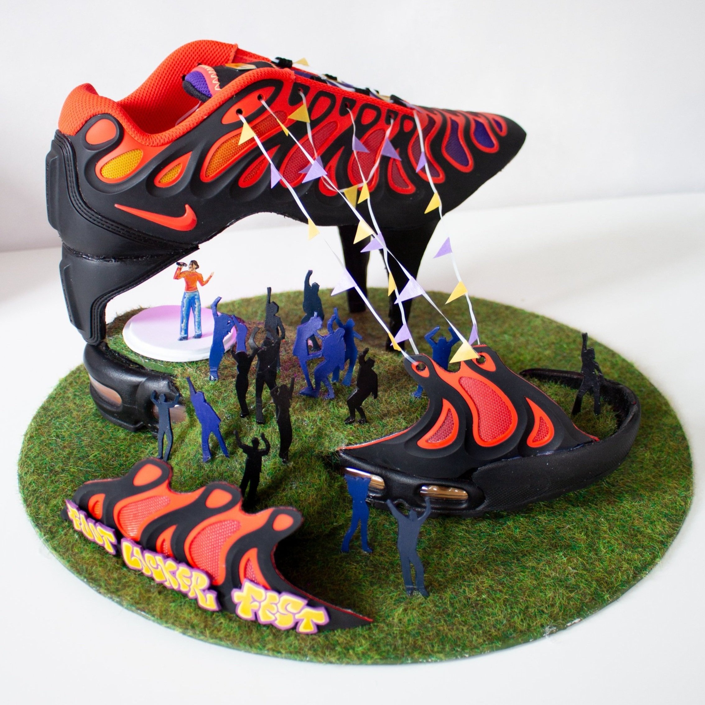 Miniature diorama created with a Nike shoe, festival theme with a stage on grass, surrounded by tiny figurines of people celebrating, with colourful bunting.