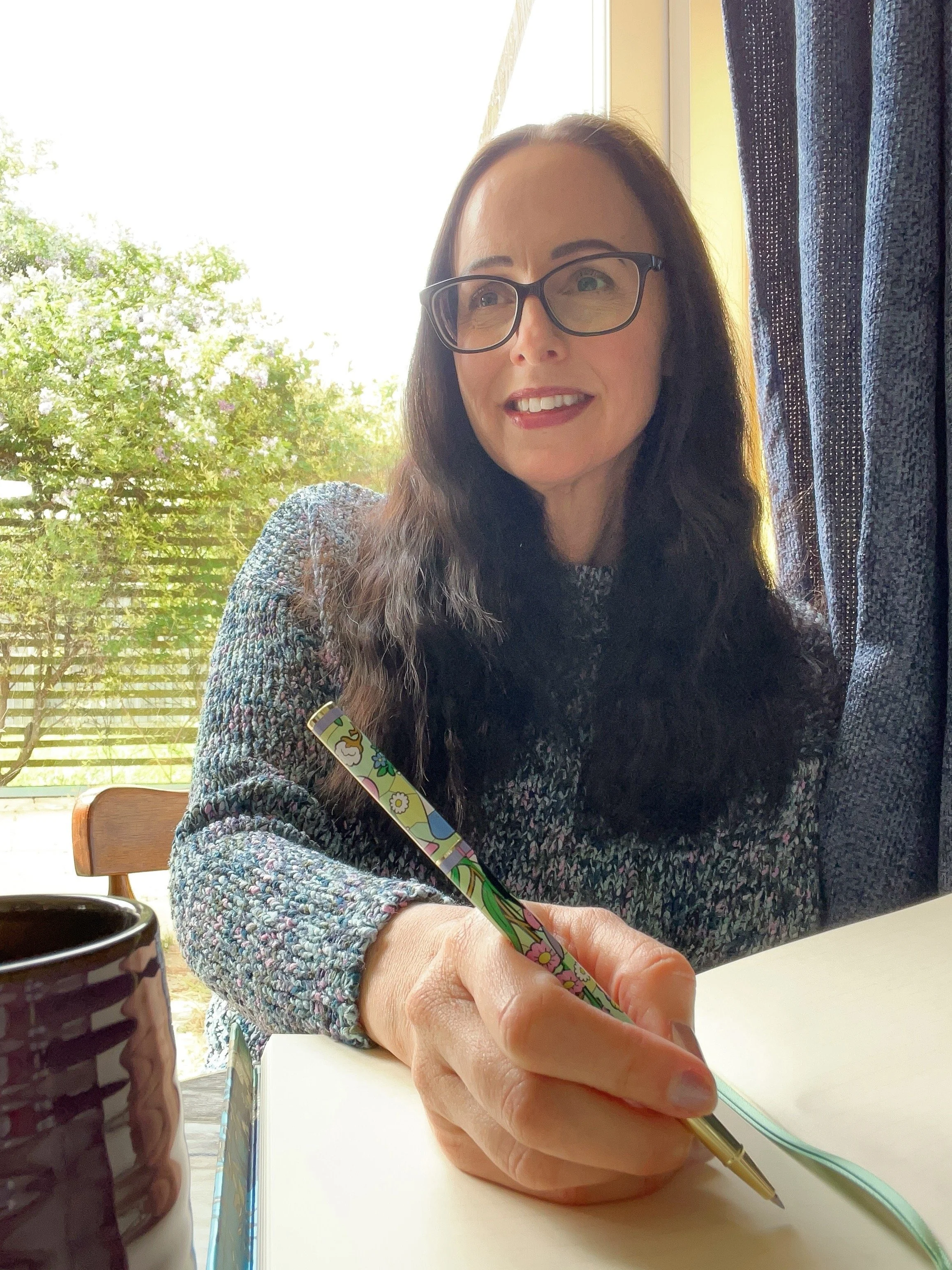 A woman with long brown hair, glasses, and a multicolored sweater sitting at a table, smiling and holding a colorful pen with a daisy and flower design. There is a mug in the foreground, and the background shows a window with trees outside.
