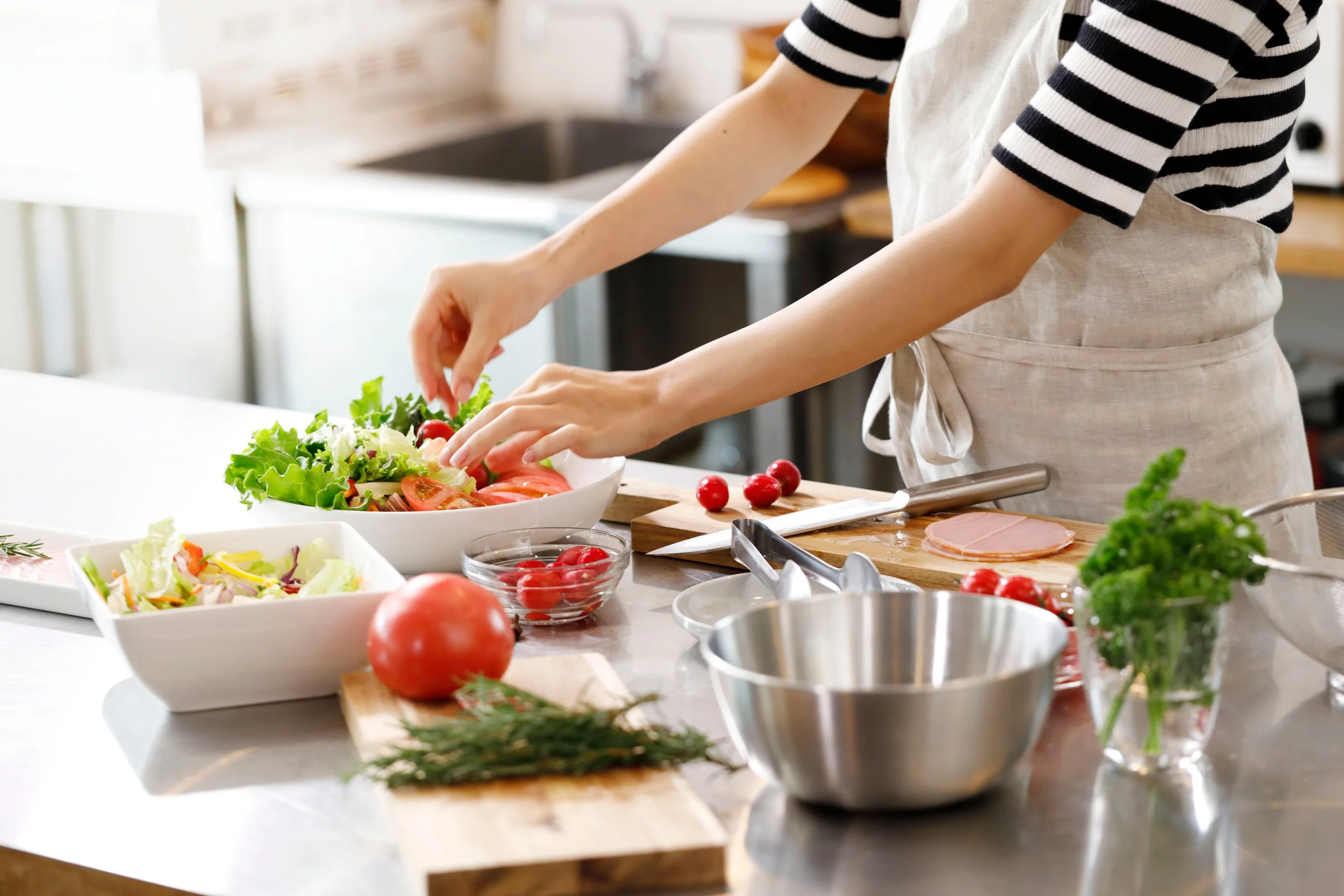 A person in a striped shirt and apron preparing a fresh salad in a kitchen, with various vegetables and bowls around.