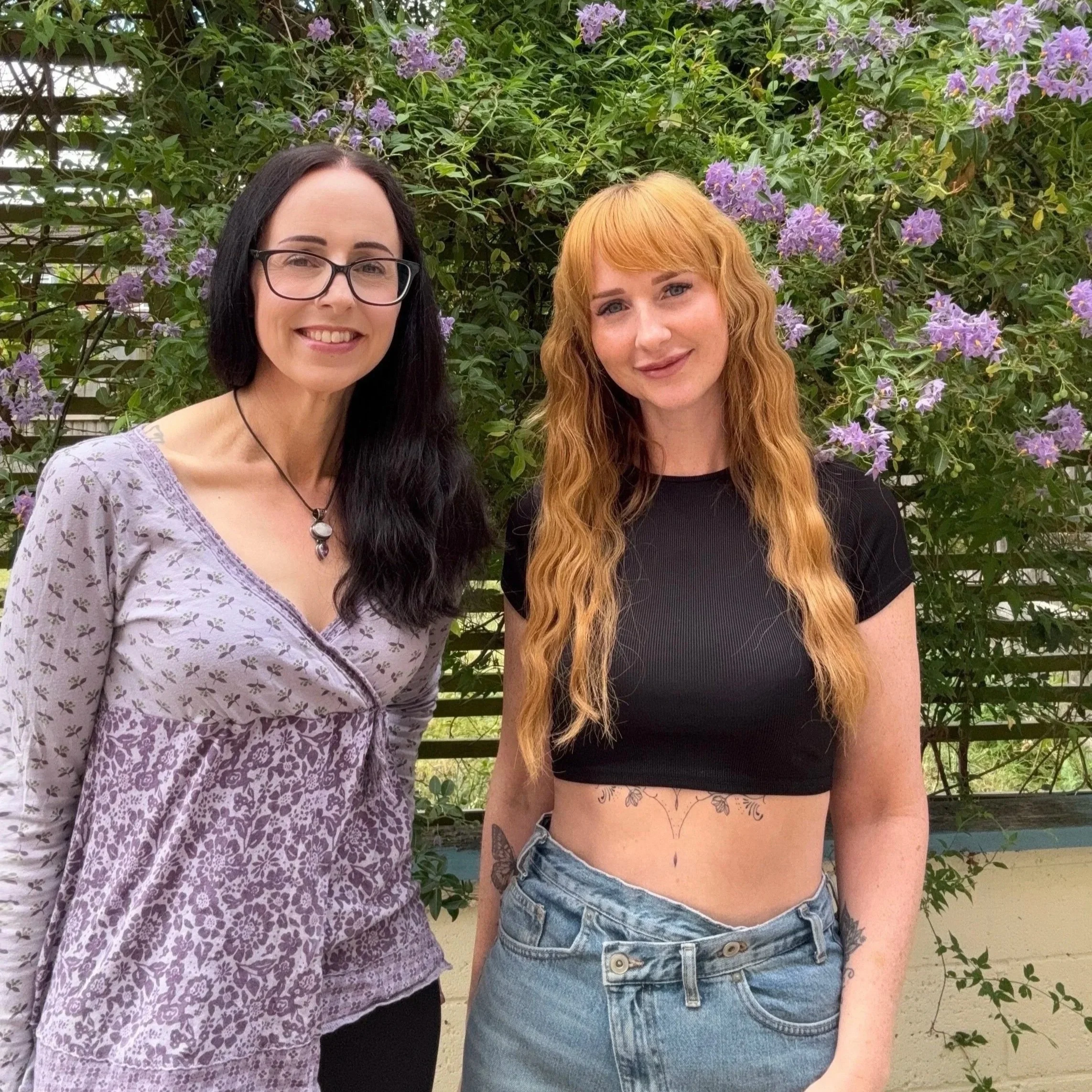 Nicky from Wright Balance Nutrition and Katie from Blossom Beauty standing together outdoors, smiling in front of a green plant wall with purple flowers.