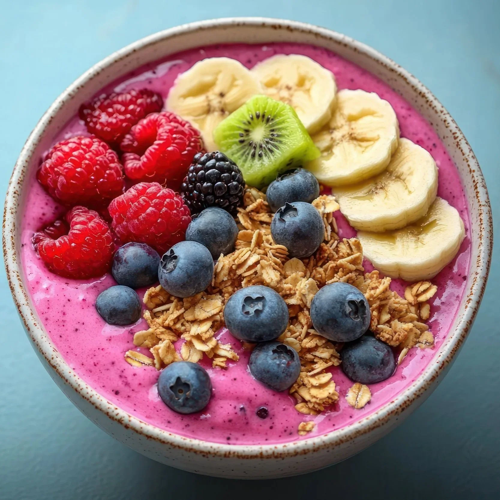 A bowl of pink smoothie topped with sliced bananas, strawberries, raspberries, blueberries, a slice of kiwi, and granola.