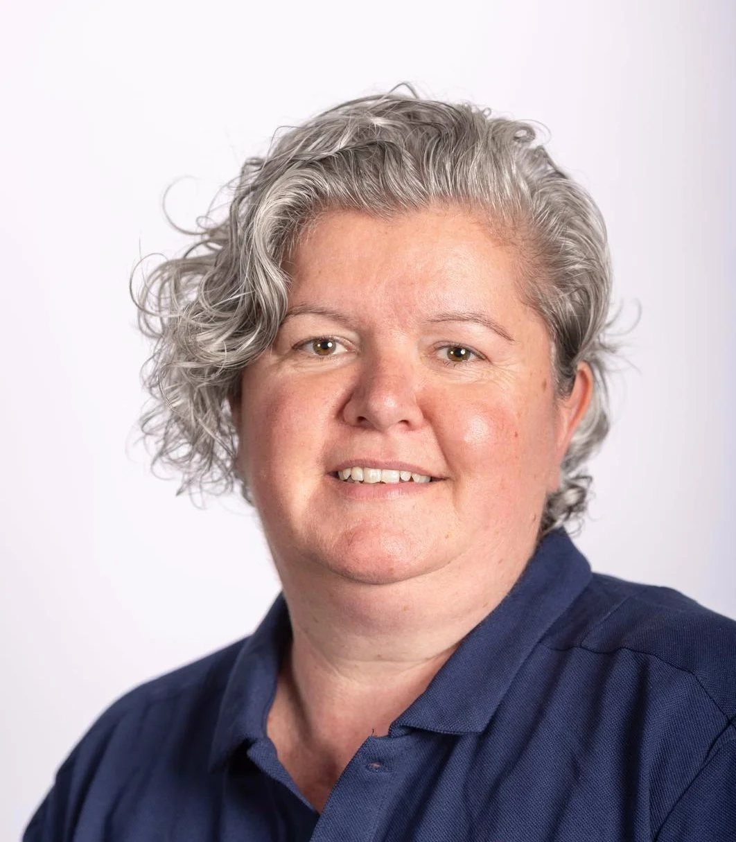 A woman with short, curly gray hair wearing a navy blue collared shirt, smiling at the camera against a plain white background.