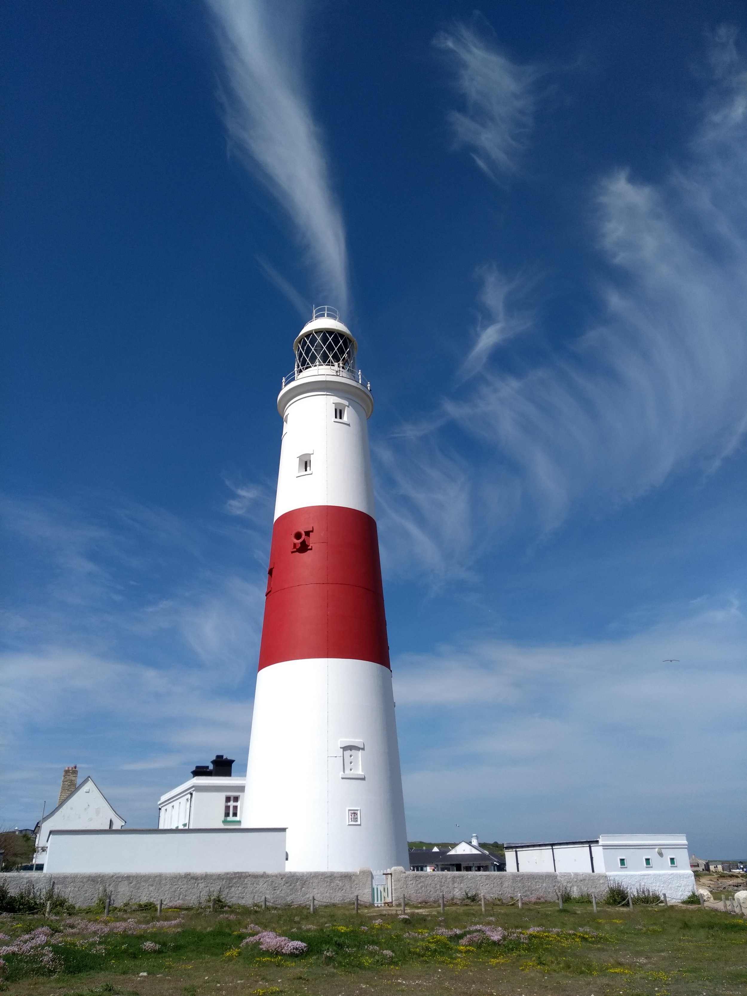 A tall white lighthouse with red horizontal stripes near the bottom, emitting a white beam of light into the clear blue sky with wispy clouds. There are some white buildings and green grass with pink and yellow flowers in the foreground.