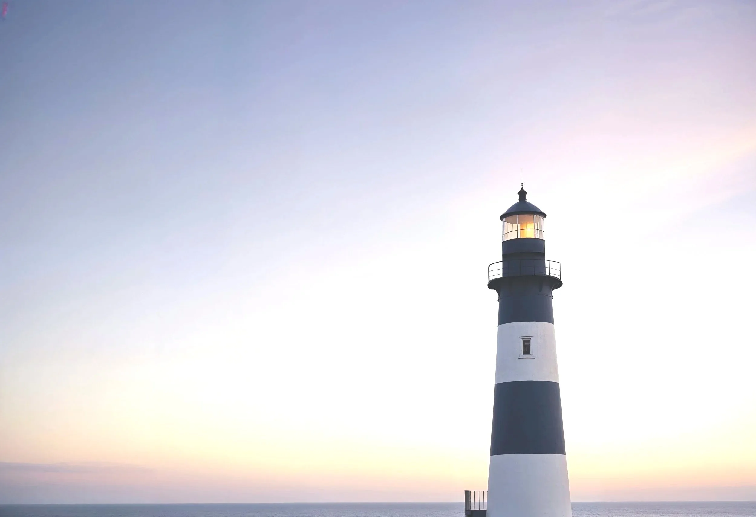 A lighthouse with black and white horizontal stripes standing by the ocean, with a clear sky during sunset or sunrise.