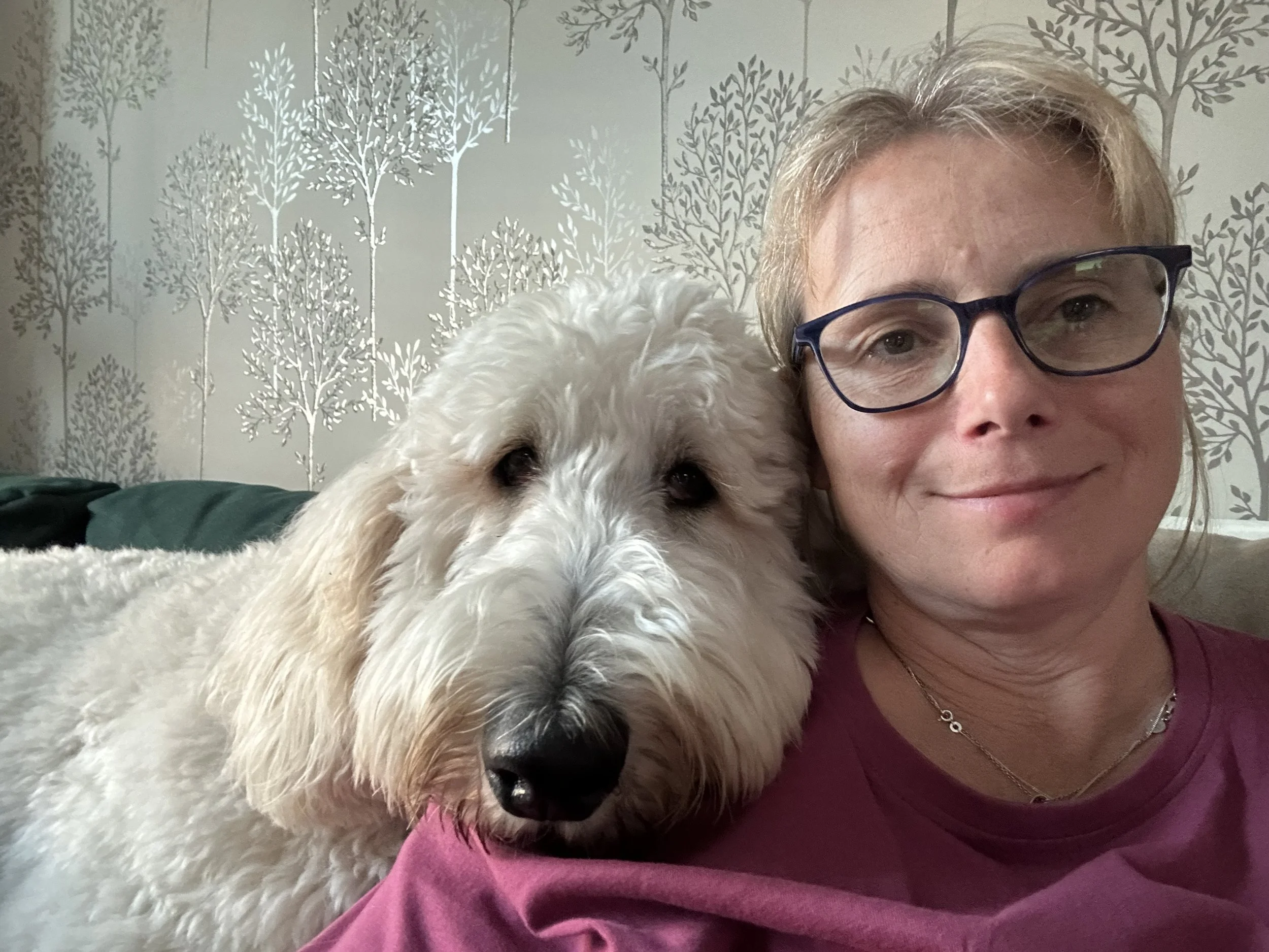 A woman with short blonde hair, glasses, and a purple shirt taking a selfie with a white curly-haired dog in front of her. The background features a wall with a gray and white tree pattern.