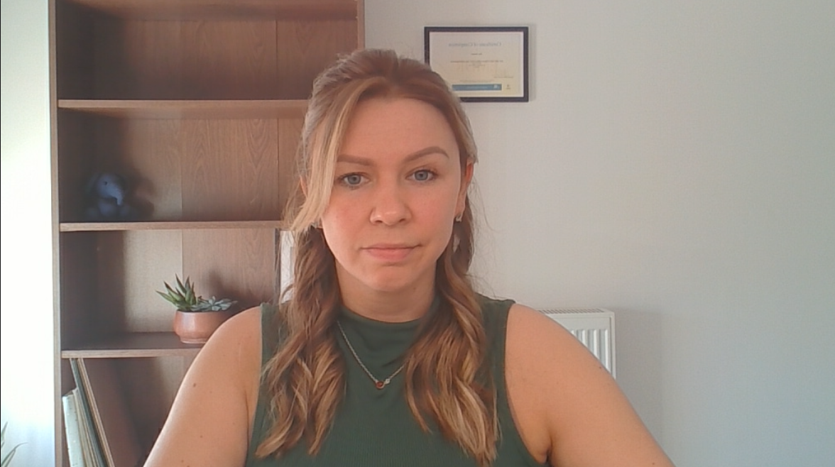 A young woman with brown hair in a half-up style, wearing a green sleeveless top, sitting indoors in an office with a bookshelf and framed certificate behind her.