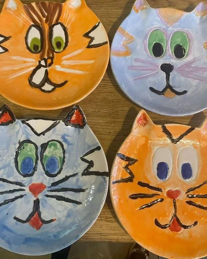 Four ceramic plates painted with cat faces in orange, white, and light purple, each with different expressions and eye colors.